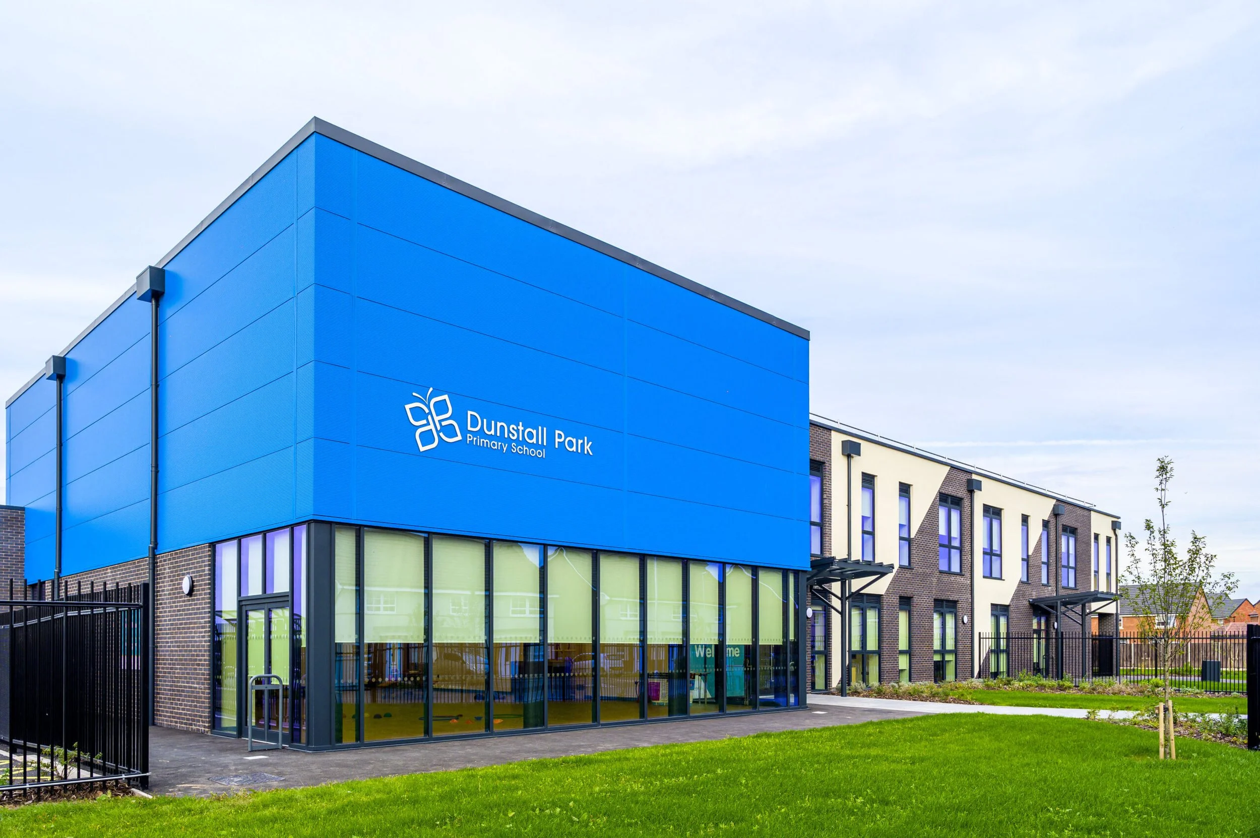 Exterior view of Dunstall Park Primary School with a blue facade, glass windows, and a green lawn.