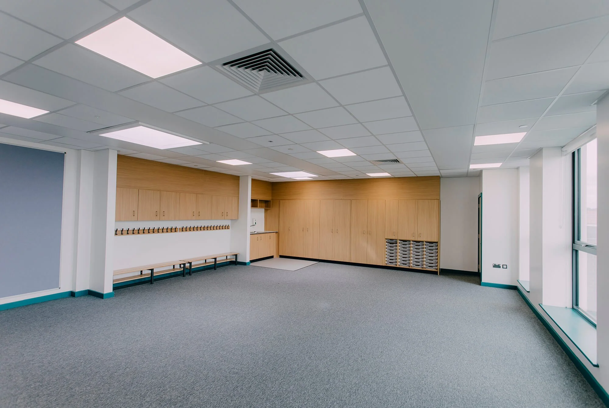 Empty office space with windows, carpeted floor, wooden cabinets, benches, and coat hooks.
