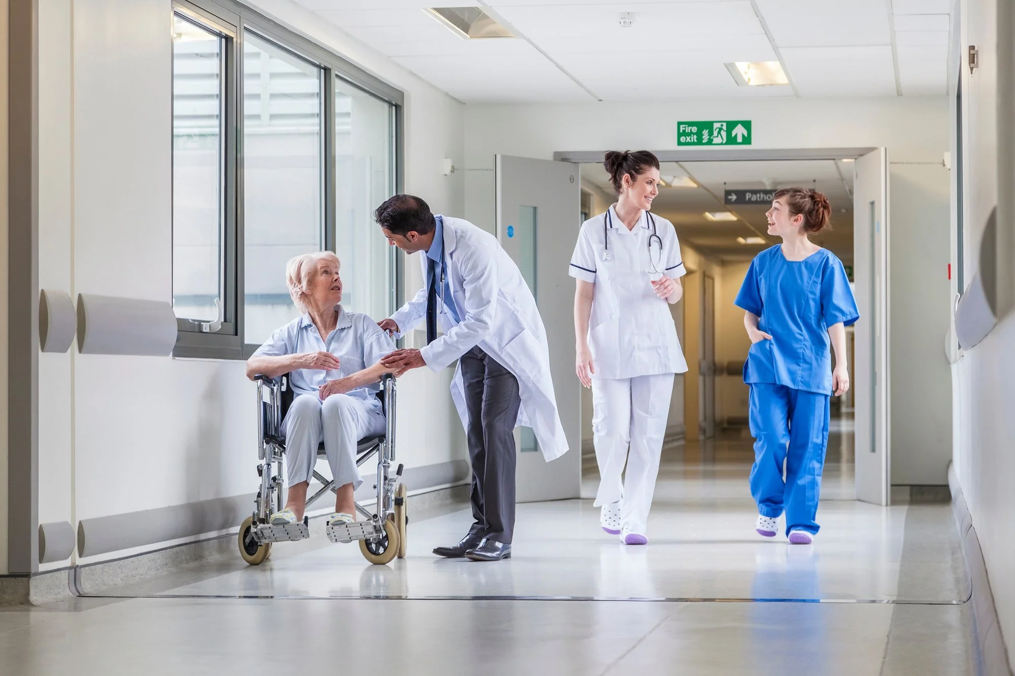 Hospital corridor with medical staff: elderly woman in a wheelchair talking to a male doctor, two nurses walking and smiling, bright environment with large windows and exit sign.