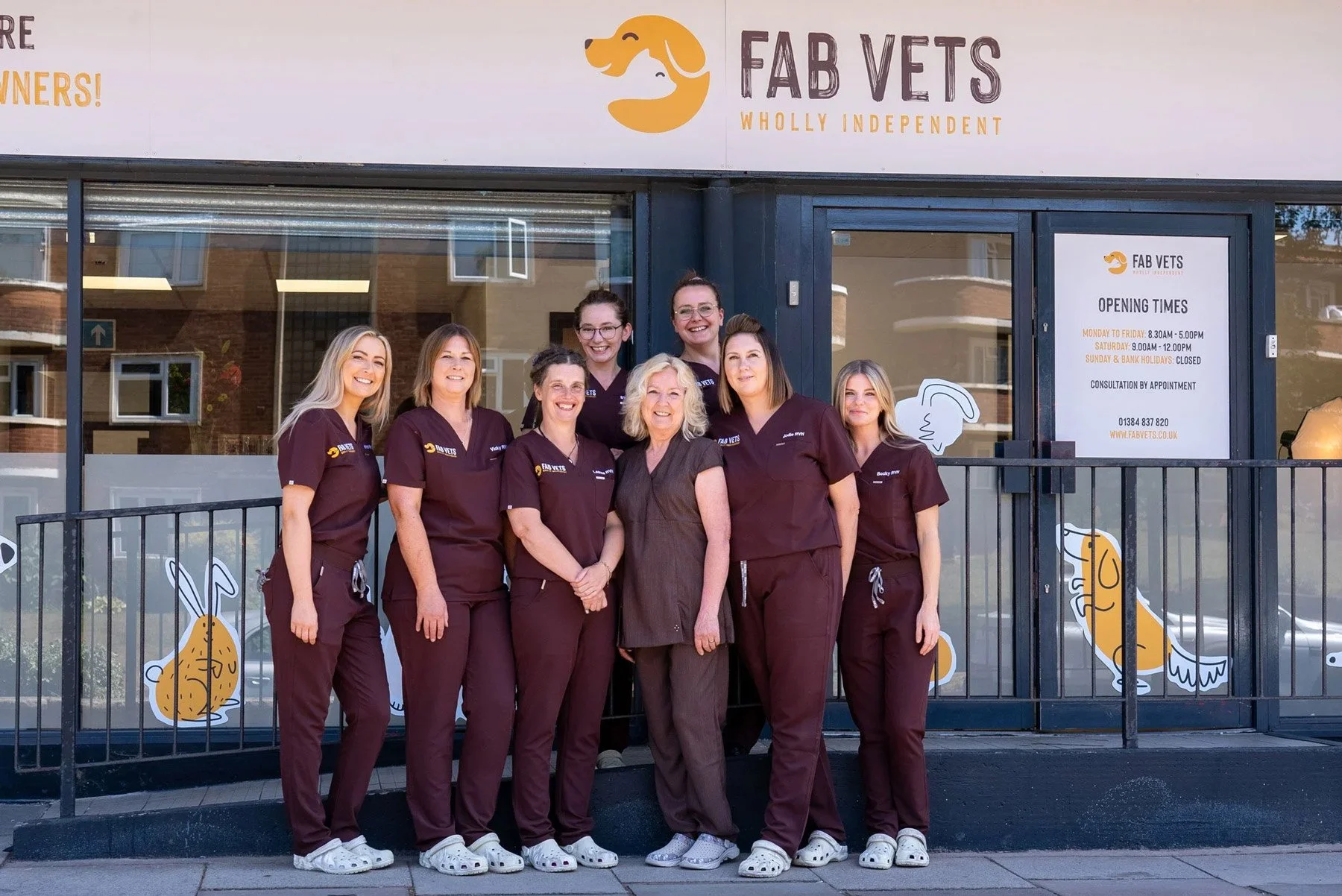 Group photo of seven women standing in front of a veterinary clinic named 'Fab Vets', wearing matching maroon uniforms, smiling at the camera.