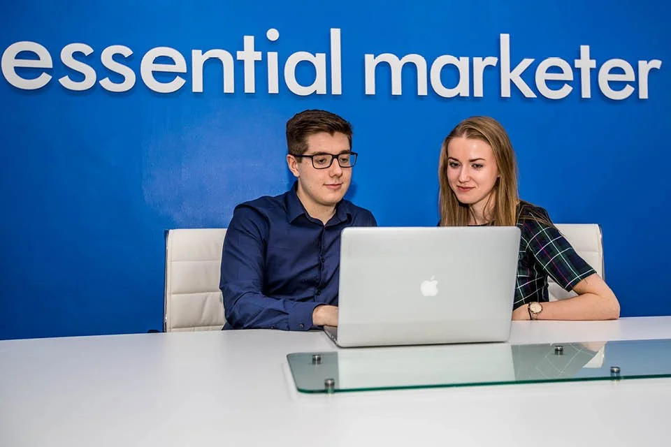 Two young professionals sitting at a desk, looking at a laptop in front of a blue wall with the words "essential marketer" written on it.