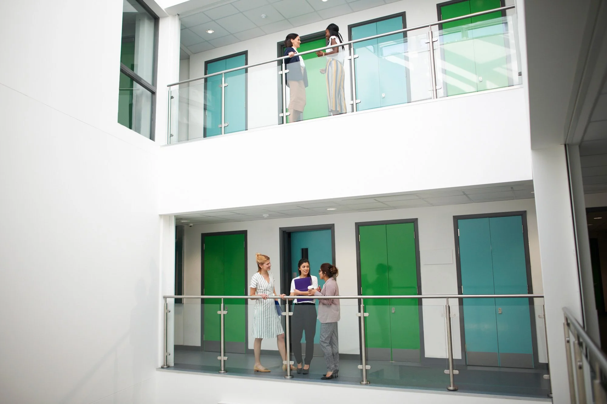 Three women are standing on a balcony of a modern office building, engaged in conversation. Two women are on the lower balcony, and one woman and another woman are on the upper balcony, talking to each other.