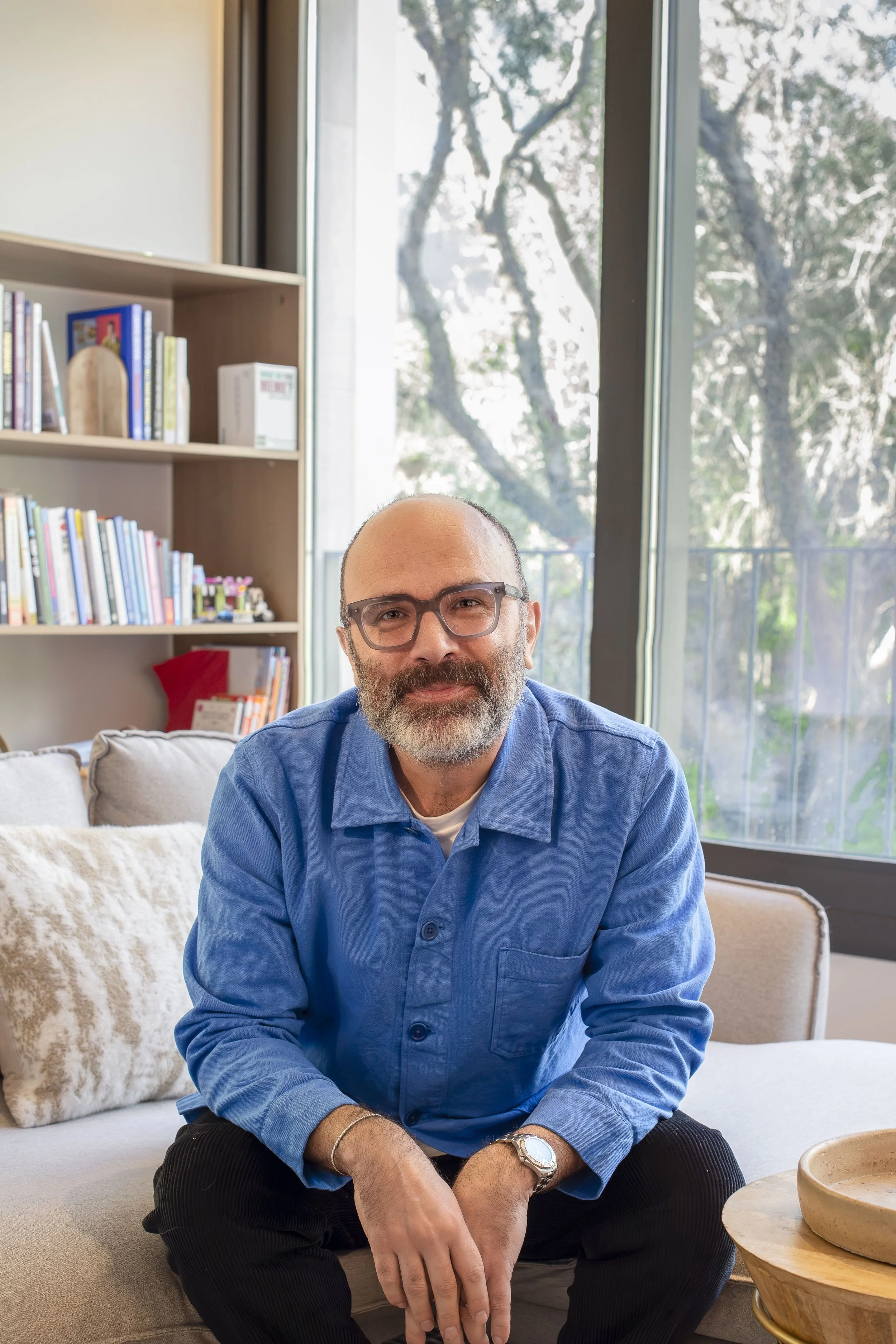 Portrait photo of a man Darran Snatchfold in a blue shirt and glasses, sitting in a room with a window behind