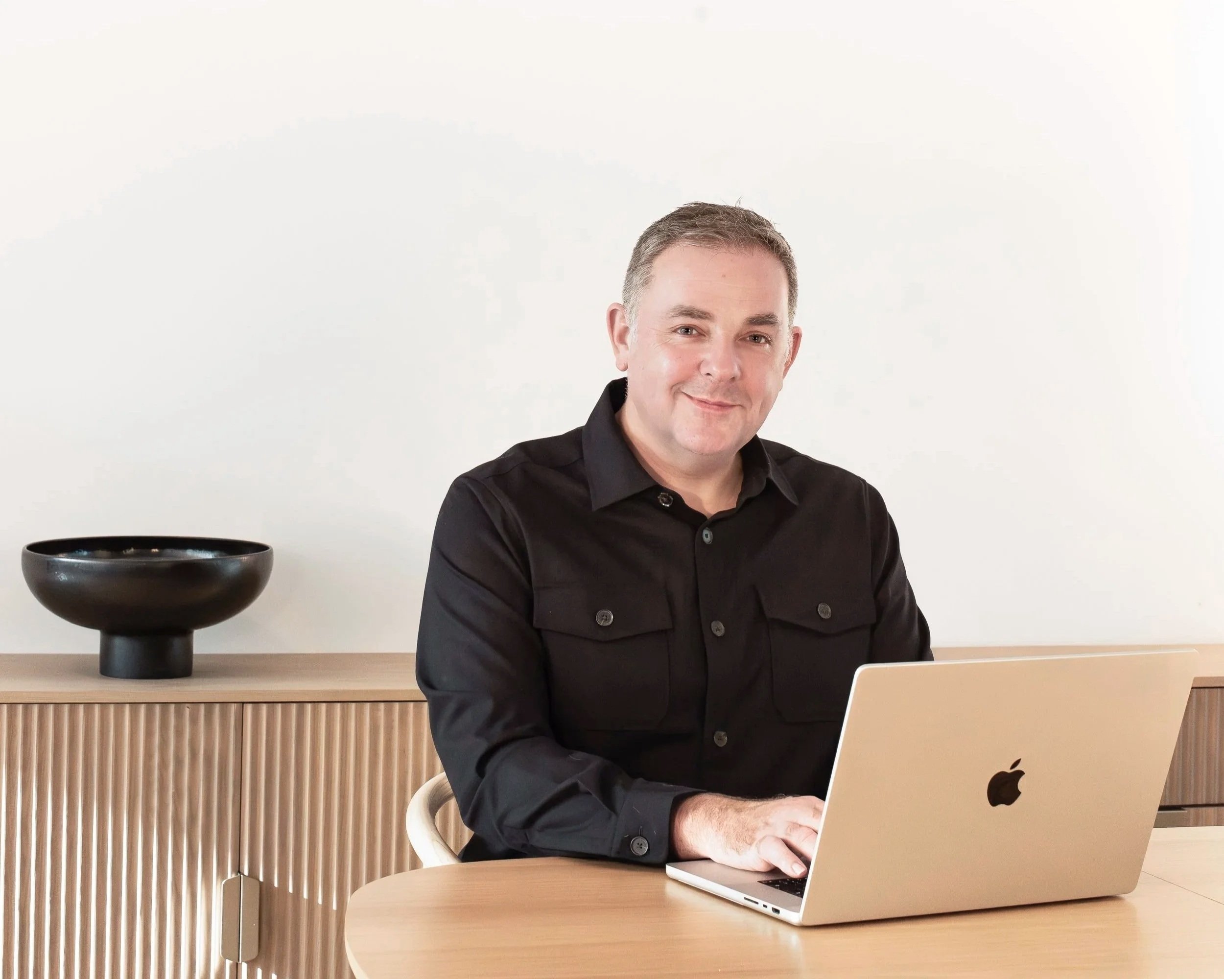 portrait of a man Alex O'Shaughnessy sitting in a room with a window behind, smiling at the camera