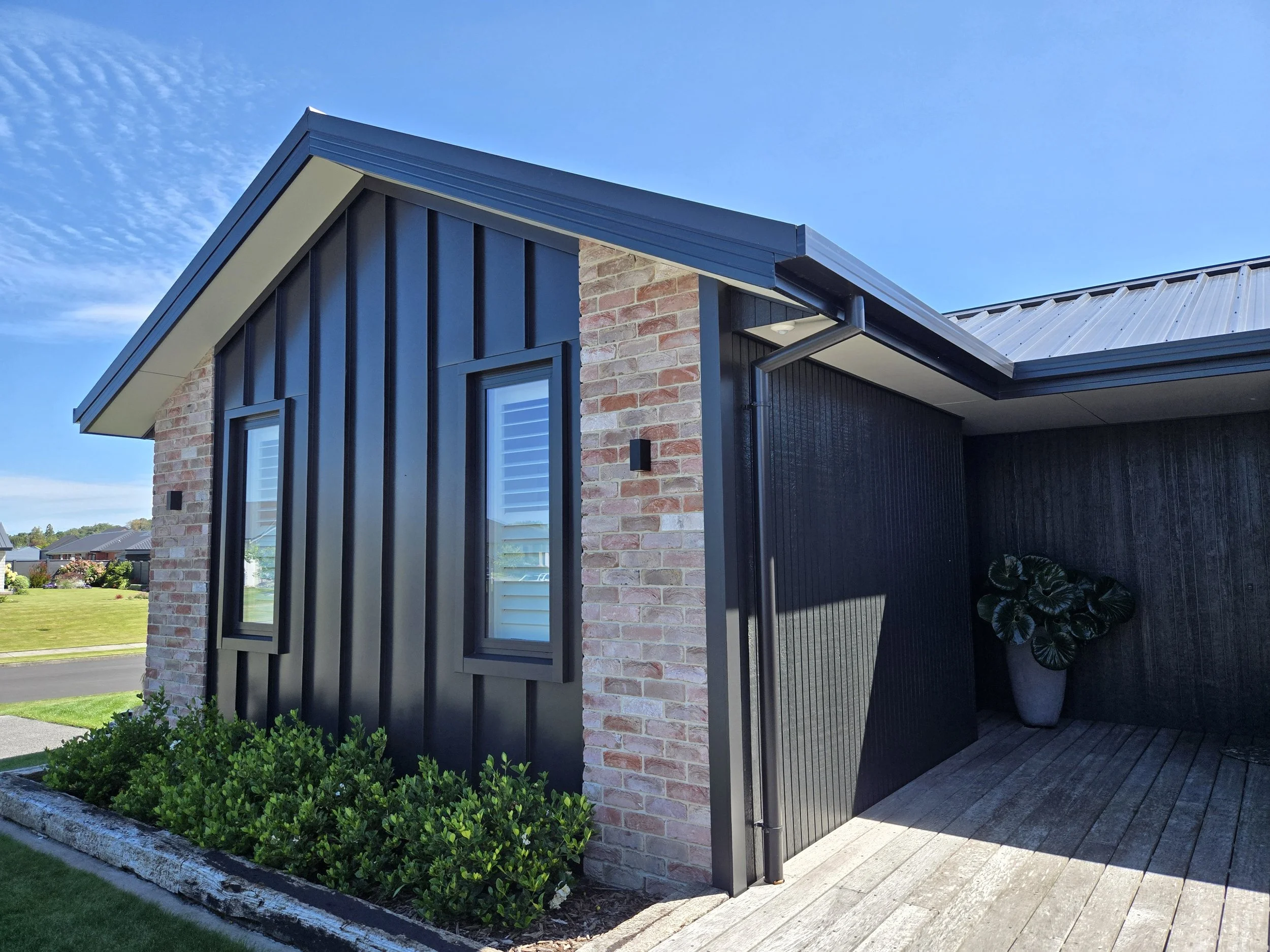 Modern house exterior with brick and black siding, small windows, and a potted plant on a wooden patio.