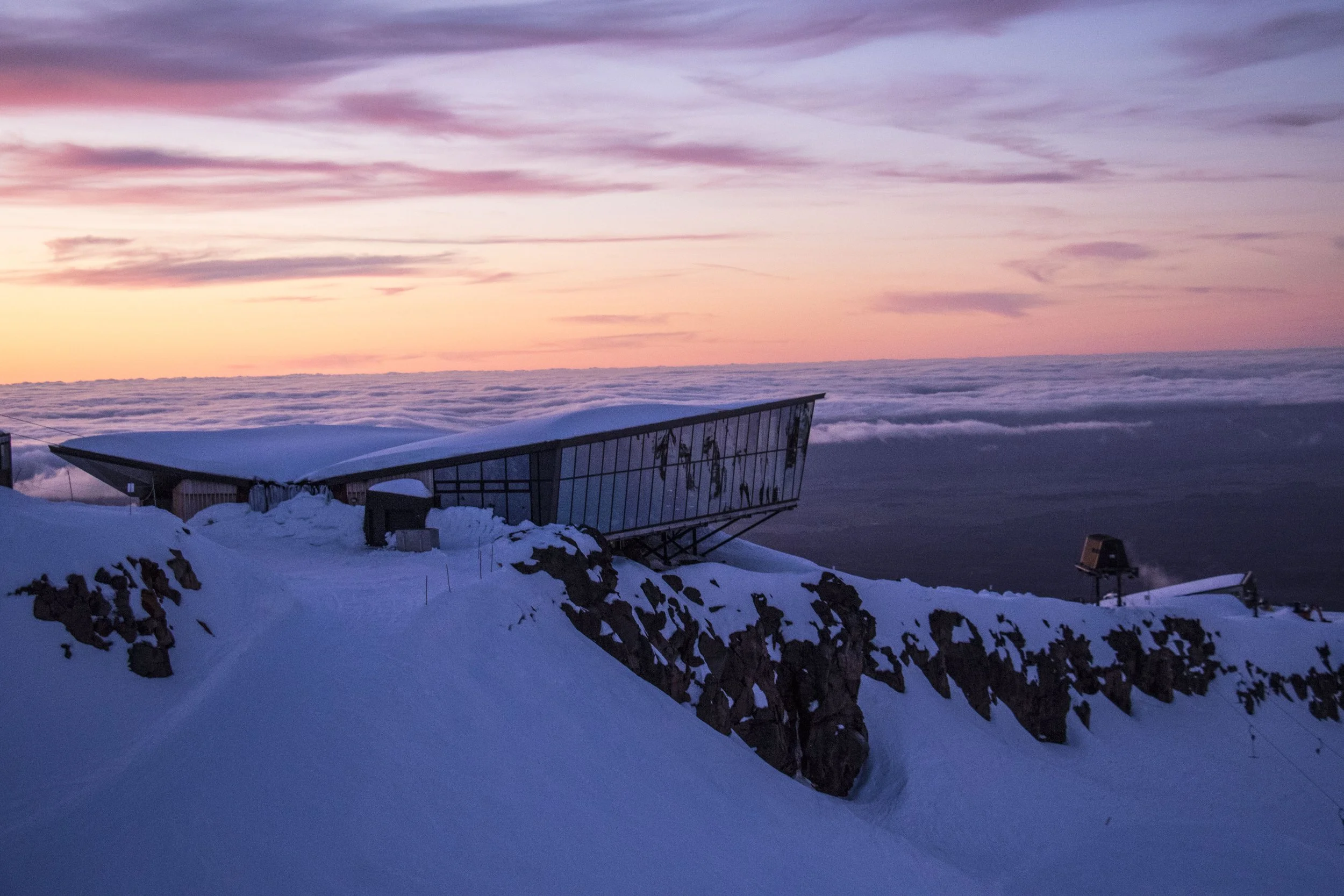Snow-covered building or structure on a mountain peak during sunset, with clouds and pinkish sky in the background.
