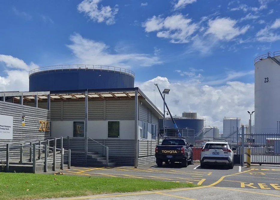 Industrial area with a building, parked cars, storage tanks, and blue sky with clouds.