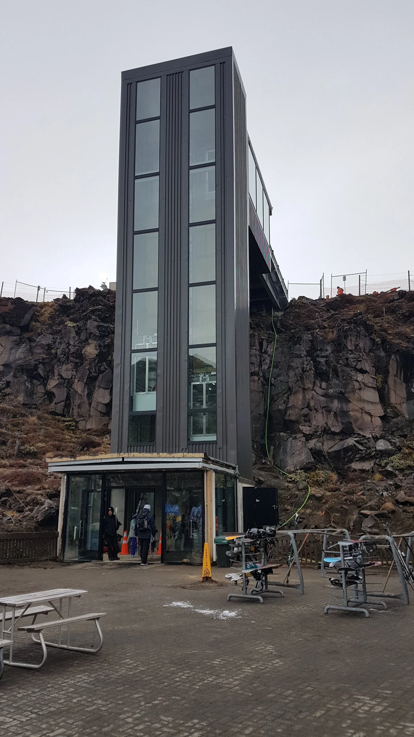 A modern glass elevator tower built into a rocky hillside with a glass door entrance at the base and multiple glass windows on the structure. There are a few people near the entrance and a bike rack in the foreground.