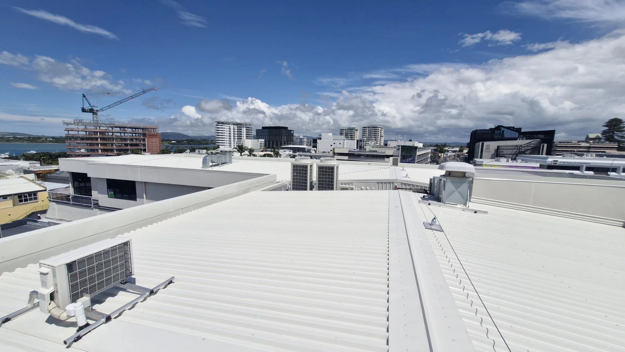 View of a city skyline from a rooftop with white metal roofing, air conditioning units, and ventilation systems, under a partly cloudy sky.
