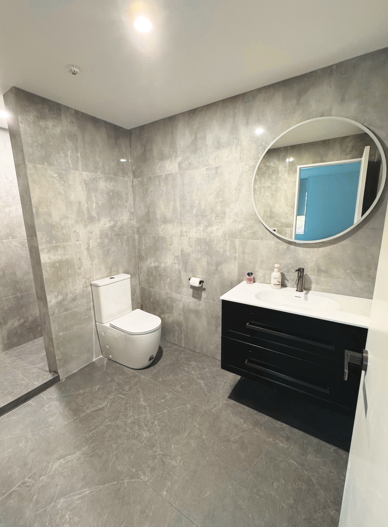 Modern bathroom with grey tiled walls and floor, a white toilet, a black vanity with a white sink, a round mirror, and hand soap and sanitizer on the countertop.