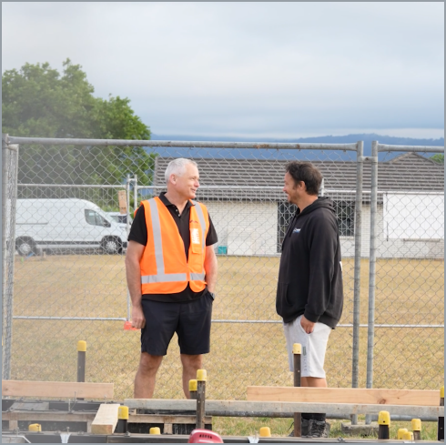Two men standing and talking in front of a chain-link fence at a construction site. One man is wearing a black shirt and shorts with an orange safety vest, and the other is wearing a black hoodie and light-colored shorts.