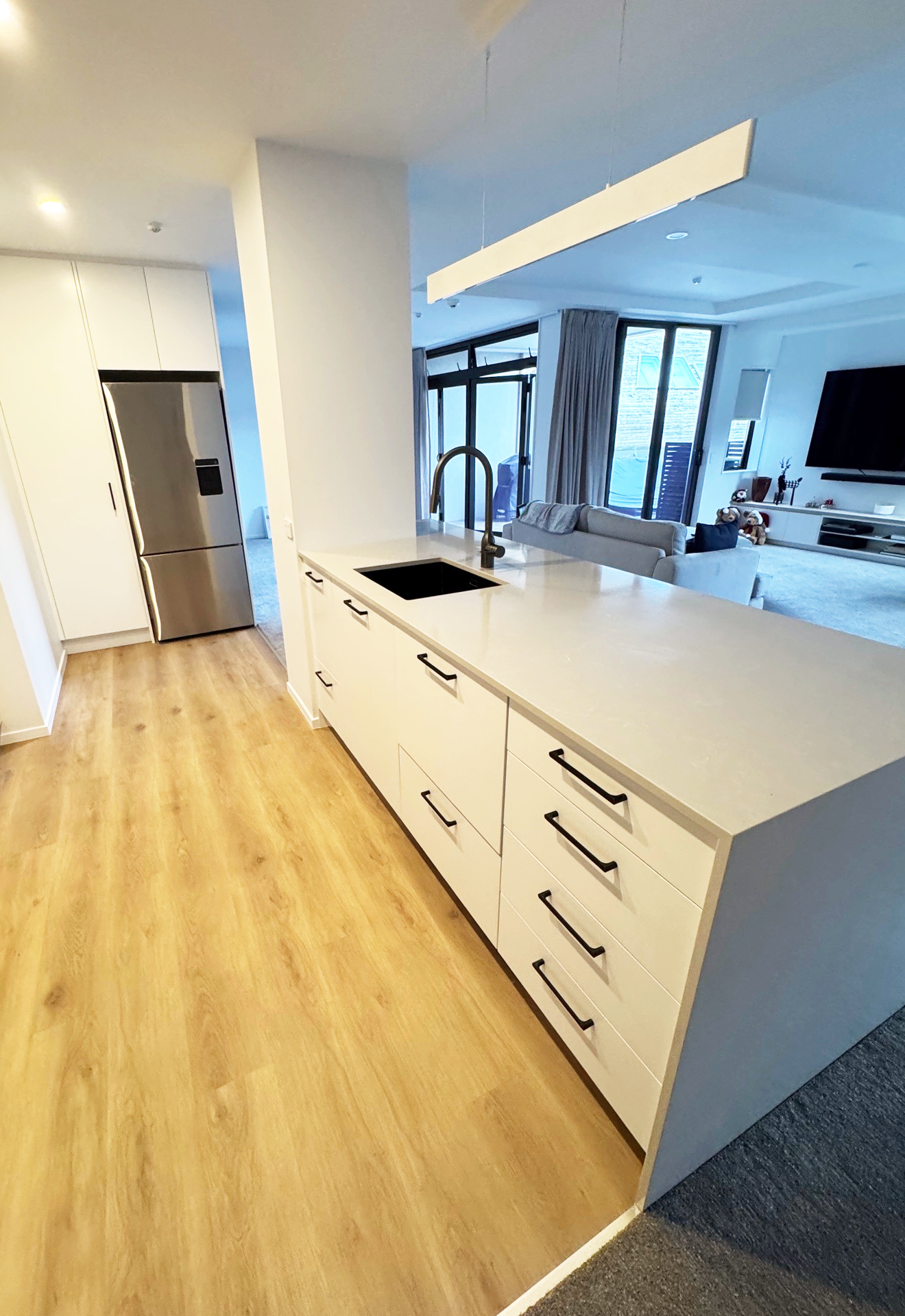 Modern kitchen with white cabinetry, black handles, light wood flooring, stainless steel refrigerator, and an open view to a living room with large windows and a TV.