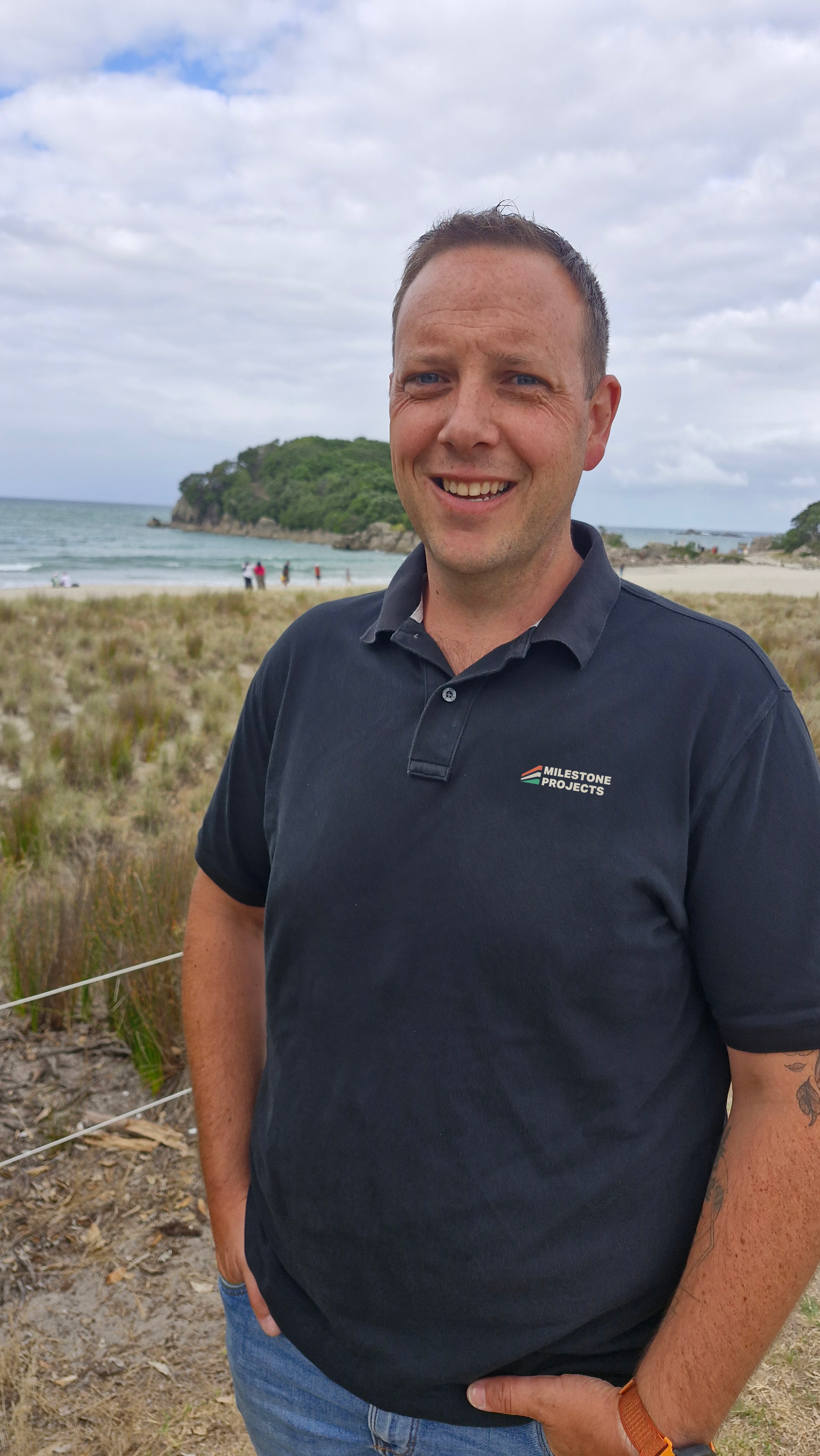A man standing outdoors on a beach, smiling, with a body of water and a small forested island in the background.