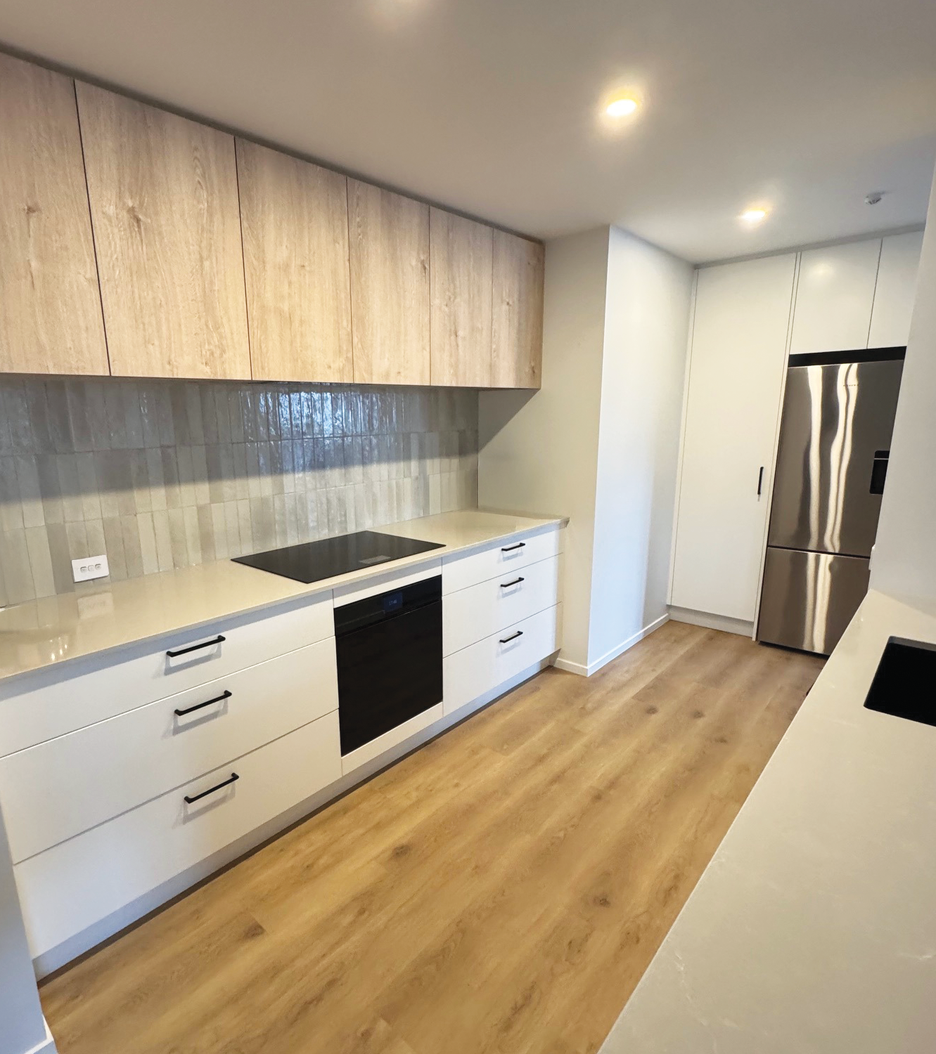 Modern kitchen with white cabinets, black handles, a black oven, wooden flooring, a beige countertop, a built-in electric stovetop, a tiled backsplash, and a stainless steel refrigerator.