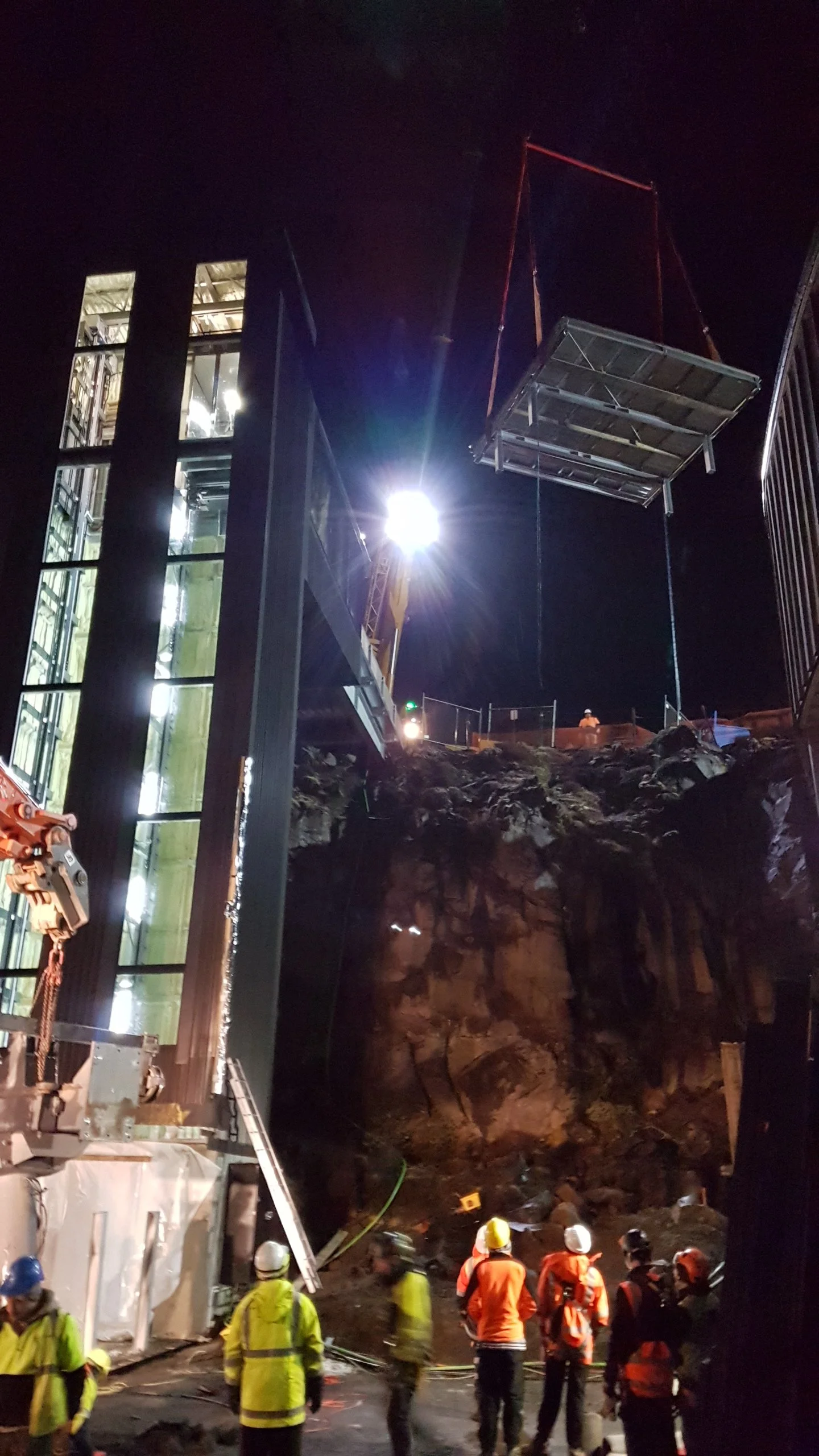 Nighttime construction site with workers wearing safety gear, a crane lifting a large platform, and a building with illuminated windows.