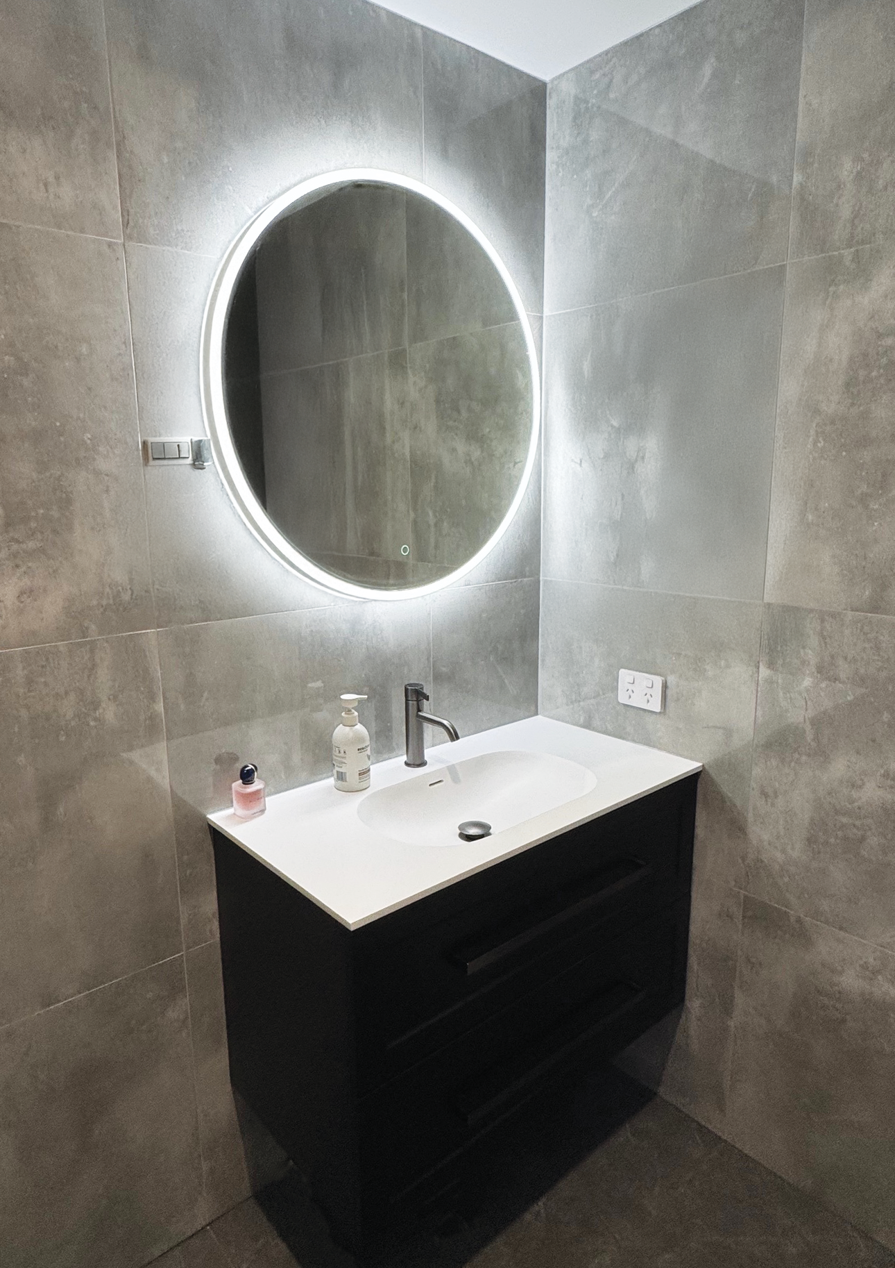 Bathroom with a round illuminated mirror, a white sink, a black vanity, a faucet, a soap dispenser, a small perfume bottle, and wall outlets.