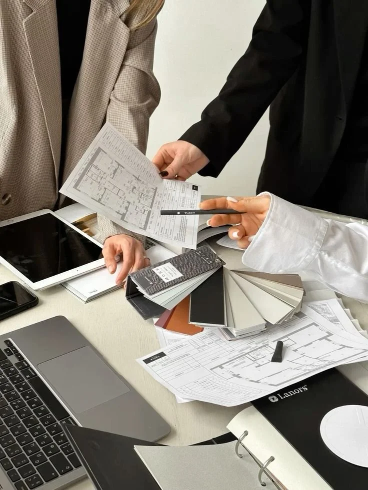 People working on interior design plans with color and material samples, floor plans, a tablet, a laptop, and a notebook on a table.