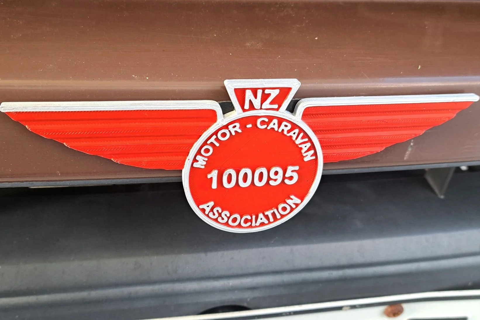 Close-up of a vintage badge with a red circular center and wings on either side, displaying the text 'NZ Motor Caravan Association' and a badge number '100095'.
