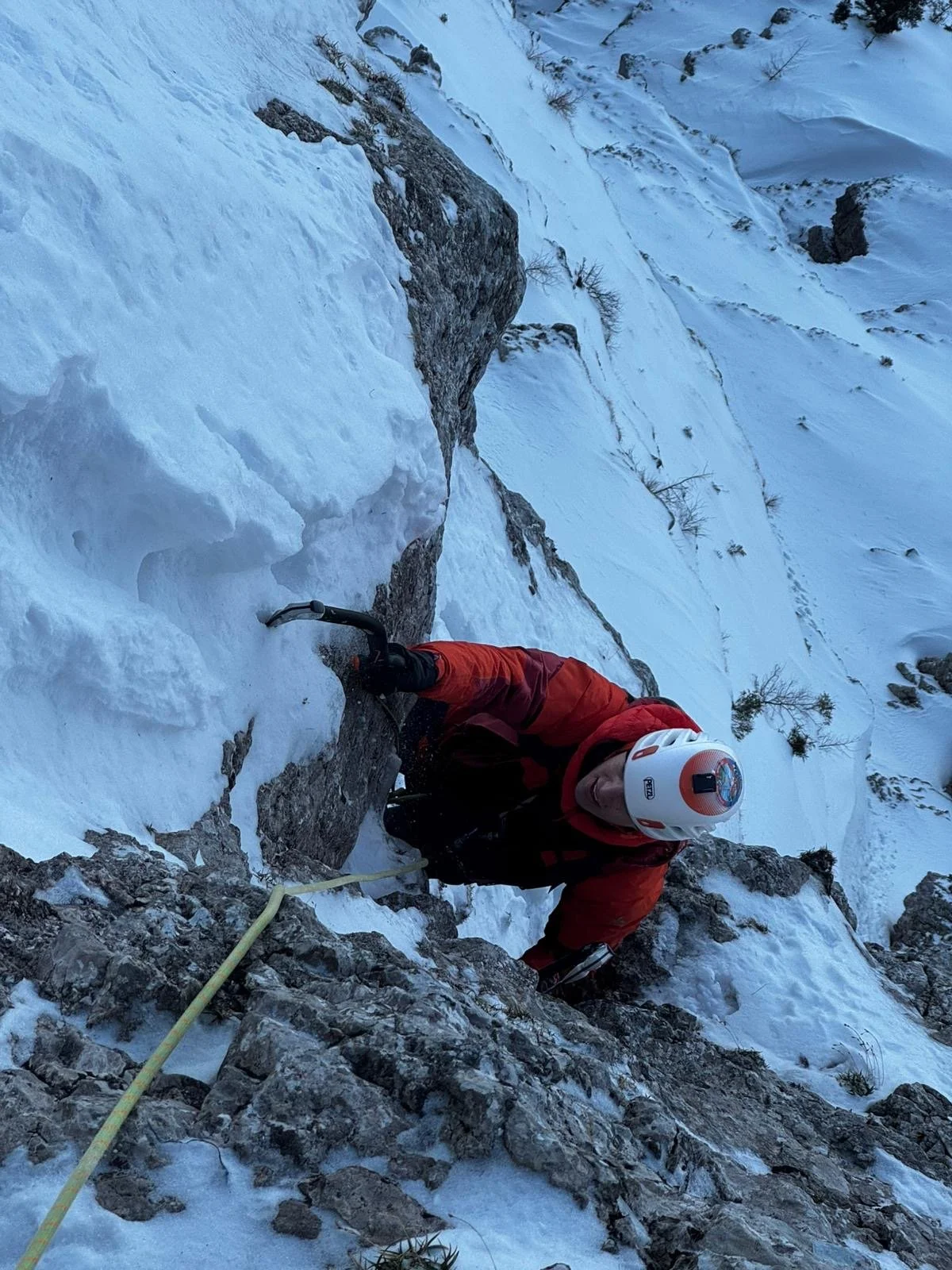 Ein Bergsteiger klettert an einer steilen, eisbedeckten Felswand mit Kletterausrüstung, einschließlich Helm und Sicherheitsseil, in einer verschneiten Berglandschaft