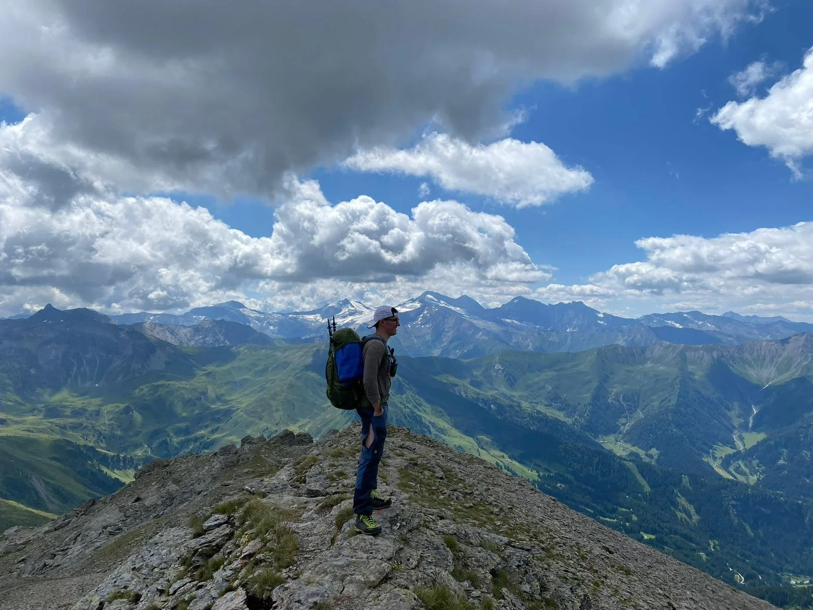 Ein männlicher Bergsteiger mit Rucksack und Sonnenbrille steht auf einem felsigen Gipfel in den Bergen, mit einer Aussicht auf grüne Tallandschaften und schneebedeckte Berge im Hintergrund, unter bewölktem Himmel.