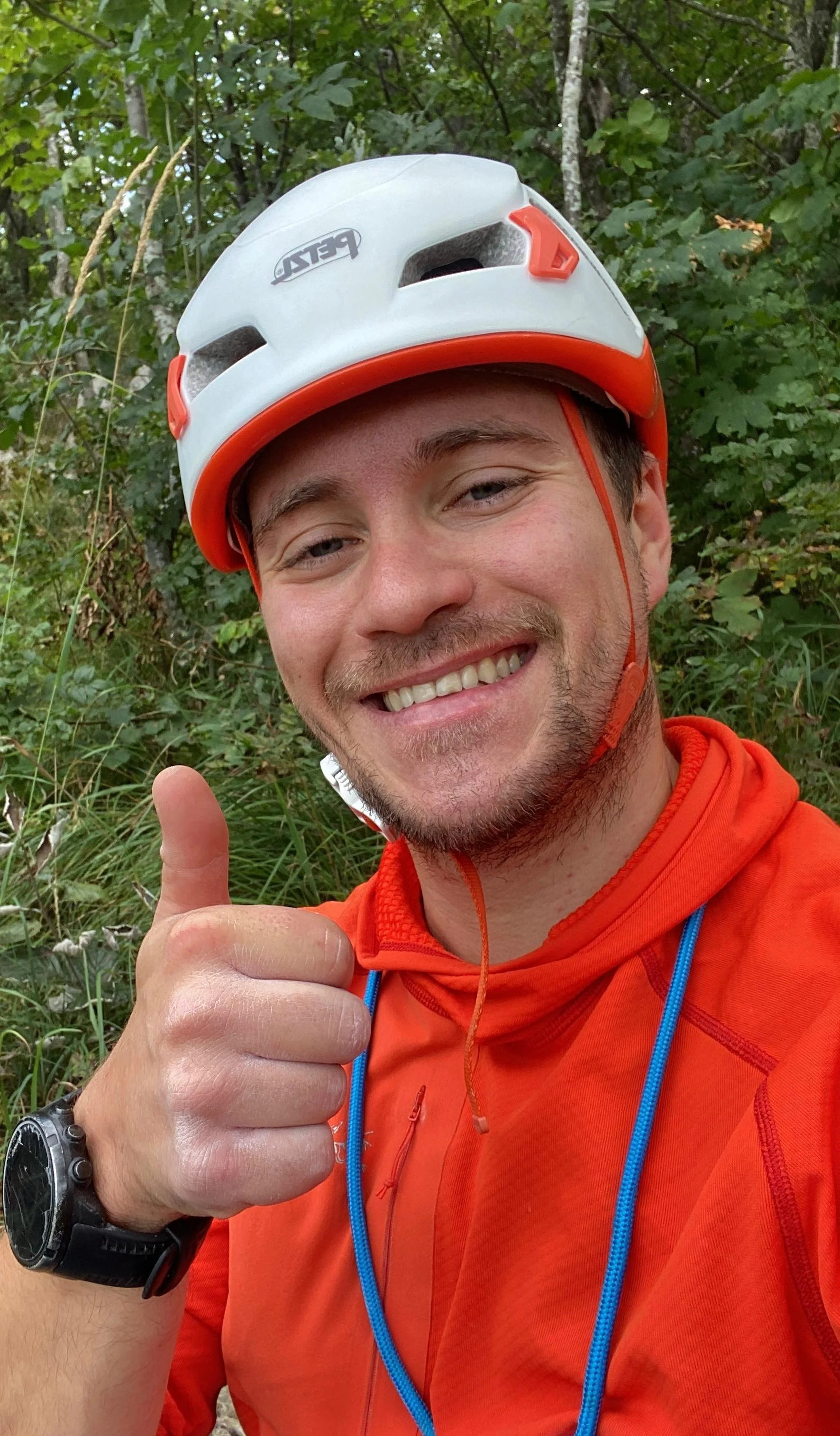 Ein lächelnder Mann trägt einen Kletterhelm und eine orange Jacke, macht ein Daumen hoch und befindet sich in einem grünen, bewachsenen Wald.