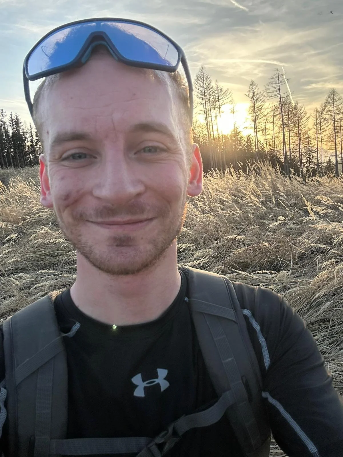 Ein Mann, der ein Selfie in einem offenen Feld bei Sonnenuntergang macht, trägt eine schwarze Sporthose und eine schwarze Unterwäsche, eine braune Rucksack sowie eine blaue Schutzbrille auf dem Kopf. Im Hintergrund sind Bäume und das gelbe Licht der untergehenden Sonne sichtbar.