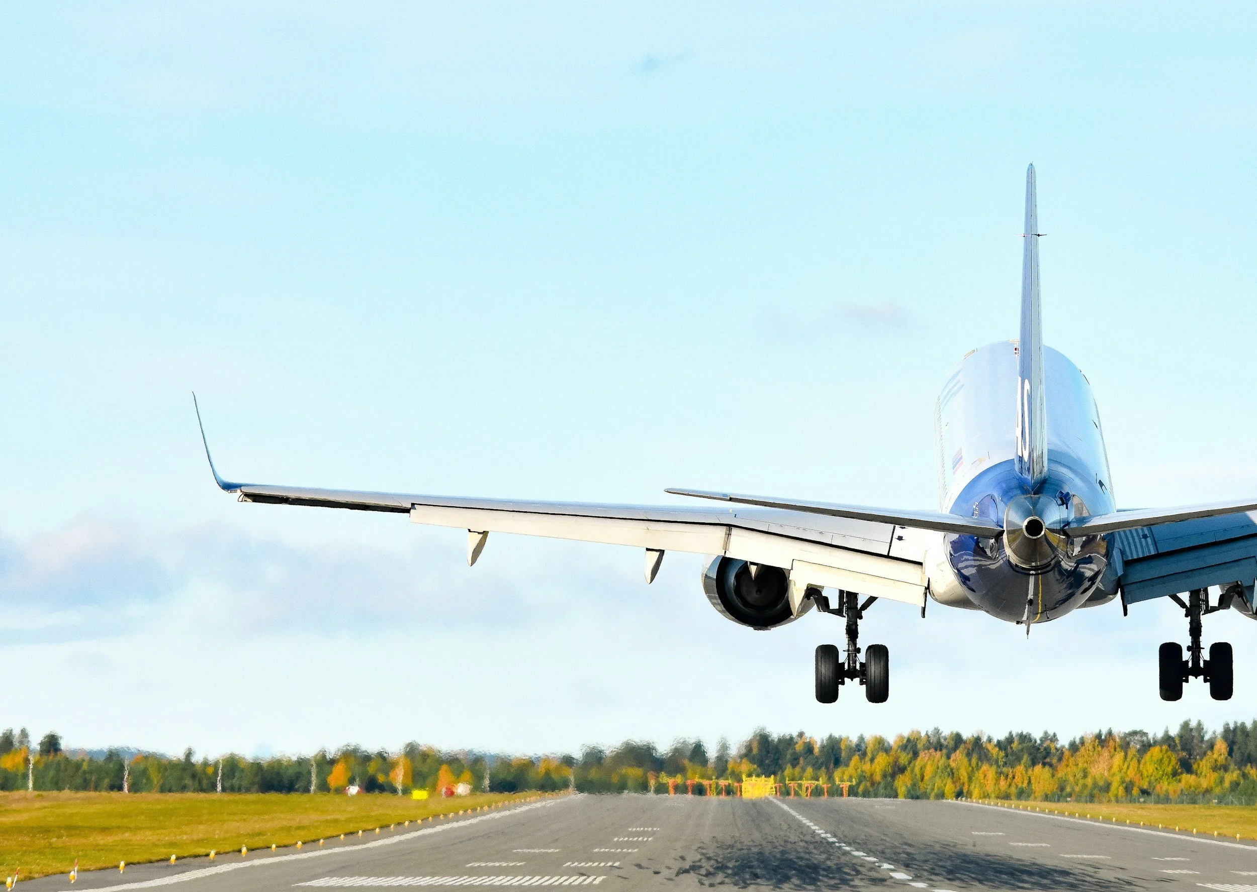 Airplane landing on runway with landing gear extended, flight path straight ahead, and blue sky overhead, colorful trees in the distance.