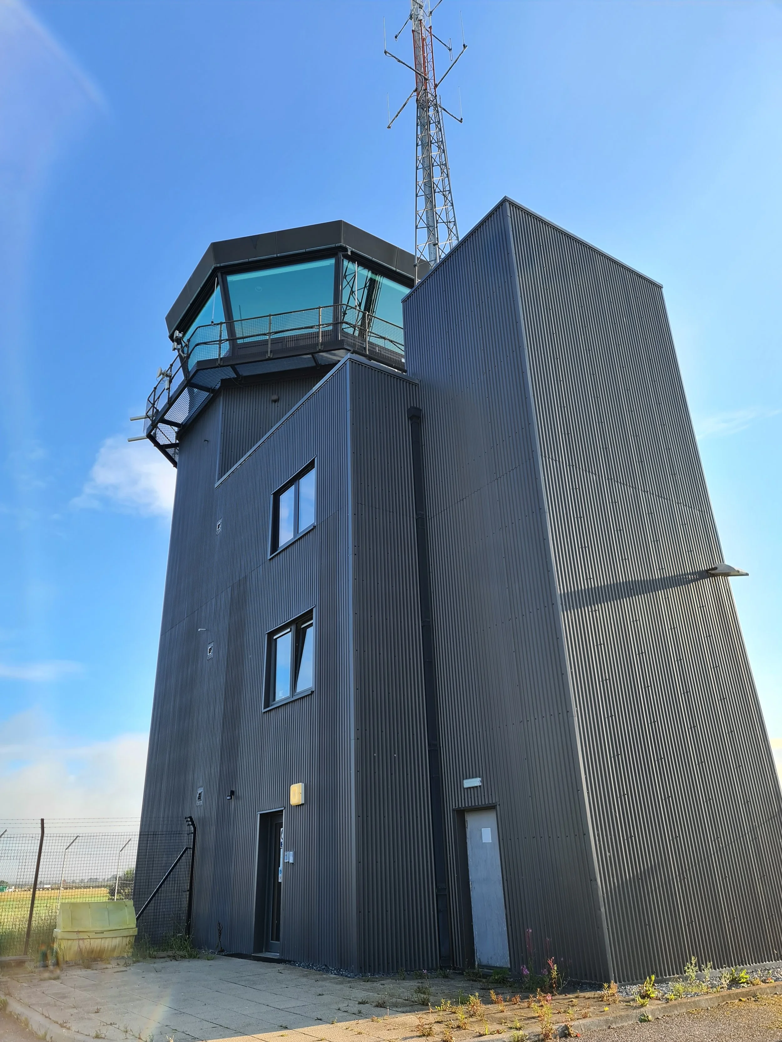 Black metal clad structure of an air traffic control tower with windows and a wrap-around balcony, topped with a tall communication antenna, against a clear blue sky.