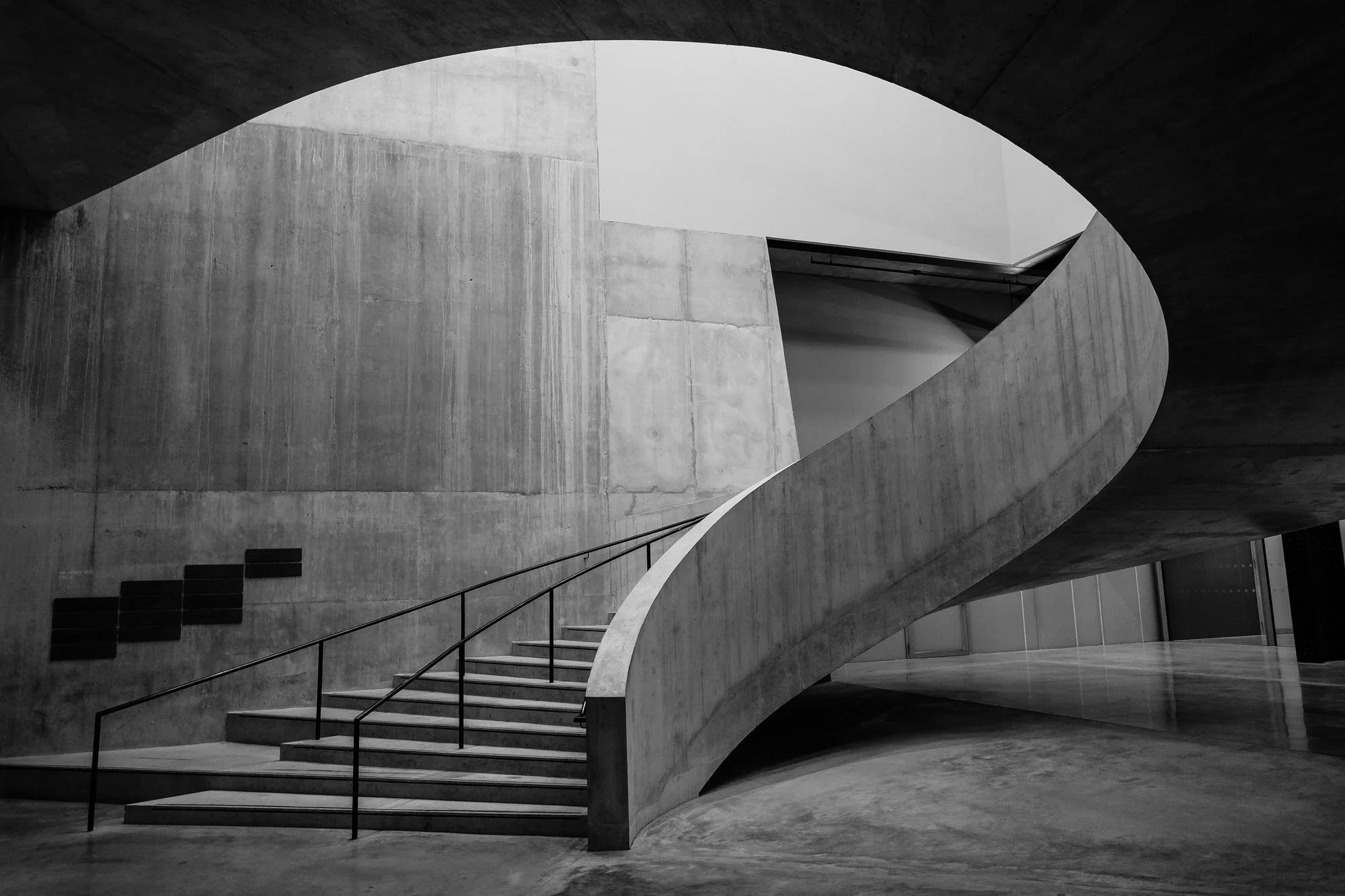 Black and white photo of a modern concrete interior featuring a spiral staircase with a curved wall and railing.