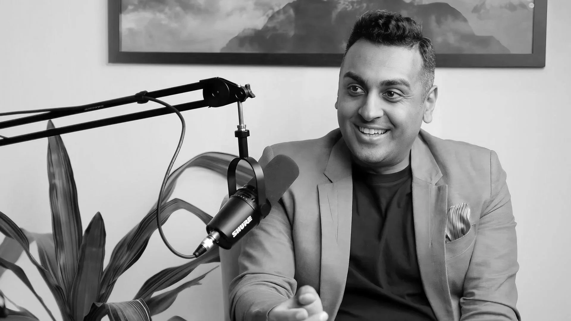 A man smiling and speaking into a microphone during a podcast or interview, with a potted plant and a framed landscape photograph in the background.