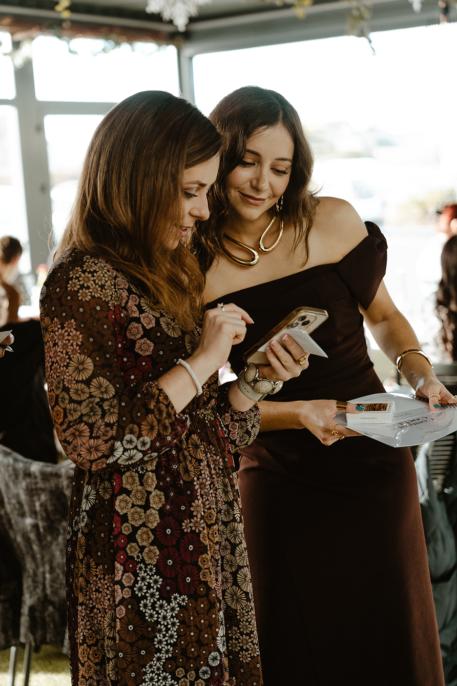 Two women are looking at a smartphone together during a social gathering. The woman on the left has long reddish-brown hair and is wearing a floral patterned dress. The woman on the right has wavy brown hair and is dressed in a dark off-shoulder dress, holding a box and dressing accessories.