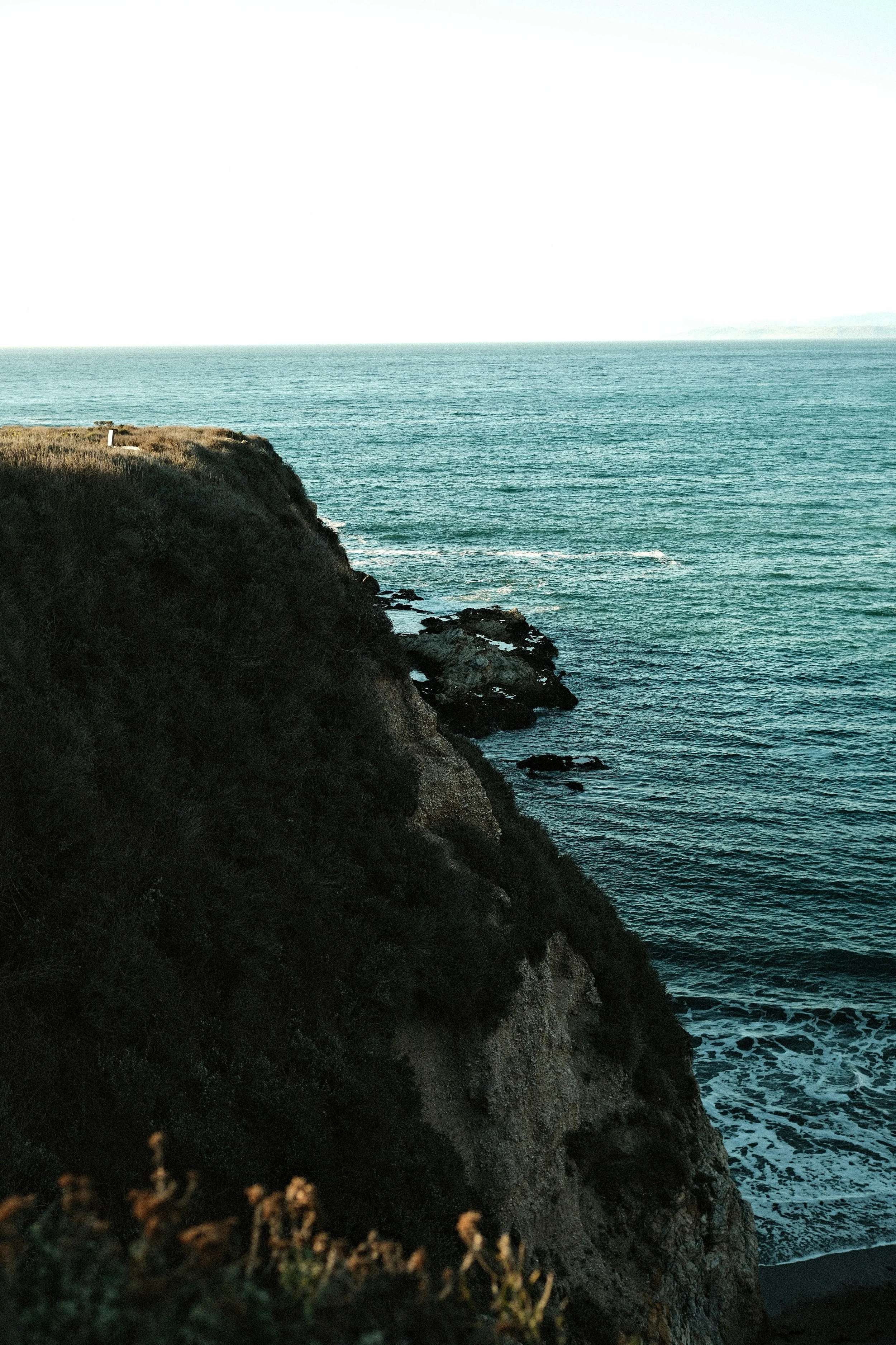 Sheep grazing on a grassy cliffside with rocks, overlooking the ocean and a rugged mountainous landscape.