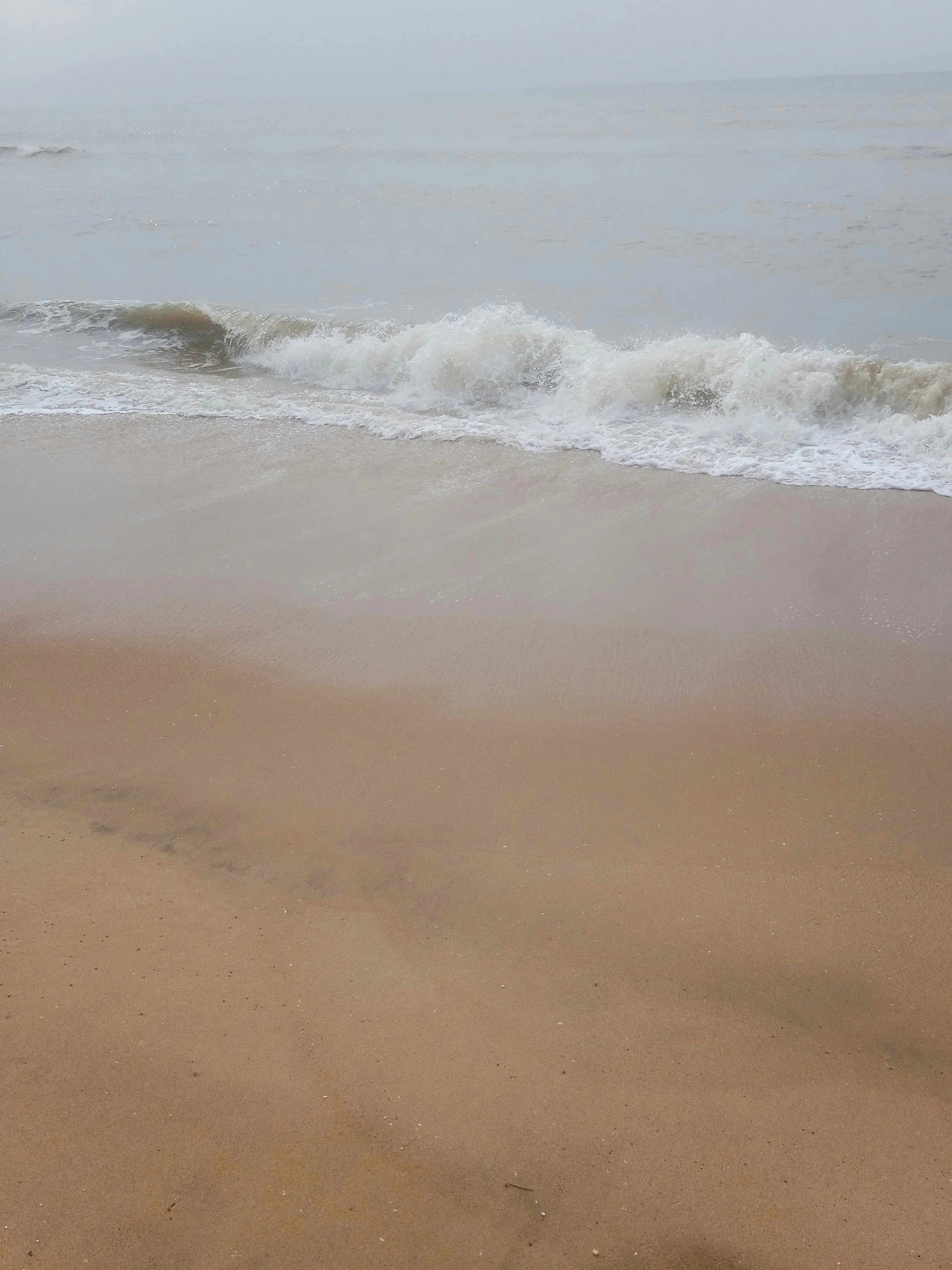 A sandy beach with rocks in the foreground and a calm ocean with small waves in the background, under a clear blue sky.