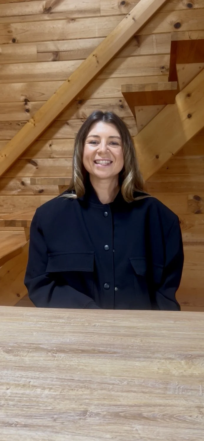 A smiling woman with shoulder-length blonde hair sitting at a wooden table in front of a wooden wall with stairs.