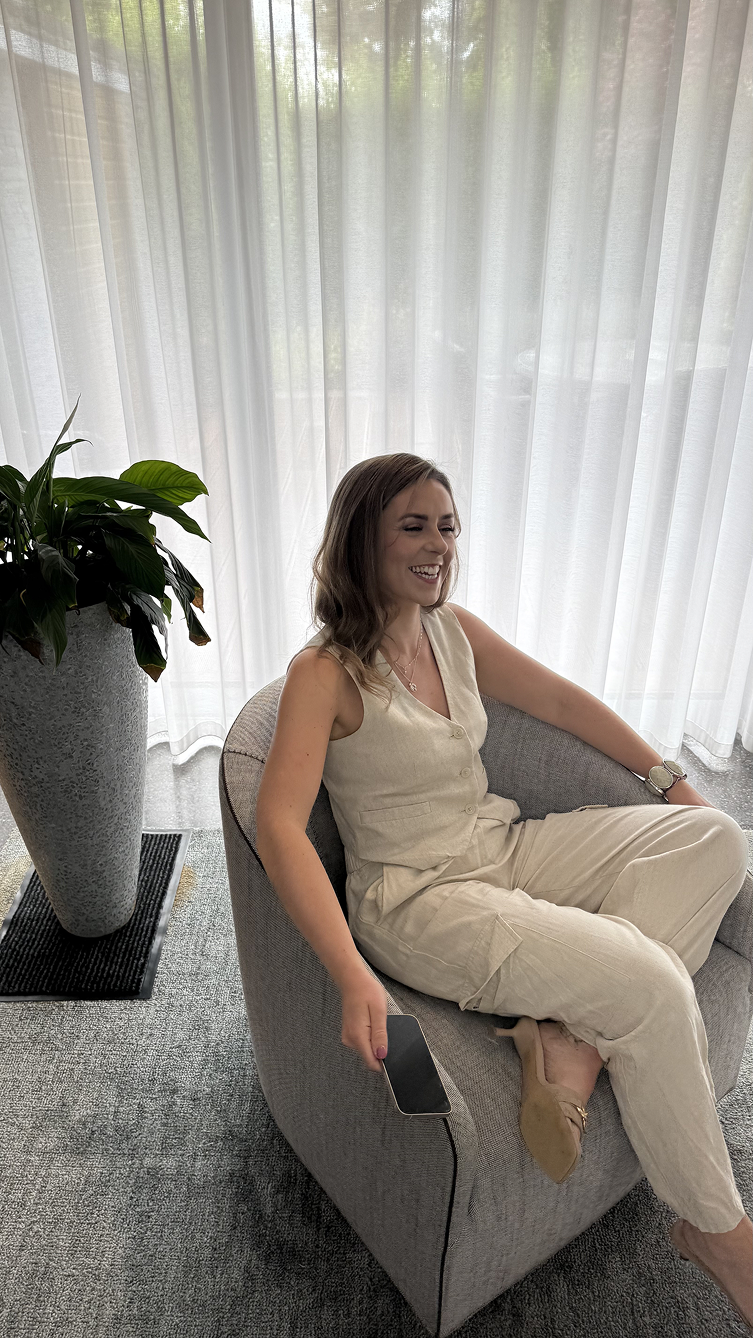 A woman with brown hair sitting on a gray armchair, smiling and holding a smartphone, next to a large potted plant near a window with sheer white curtains.