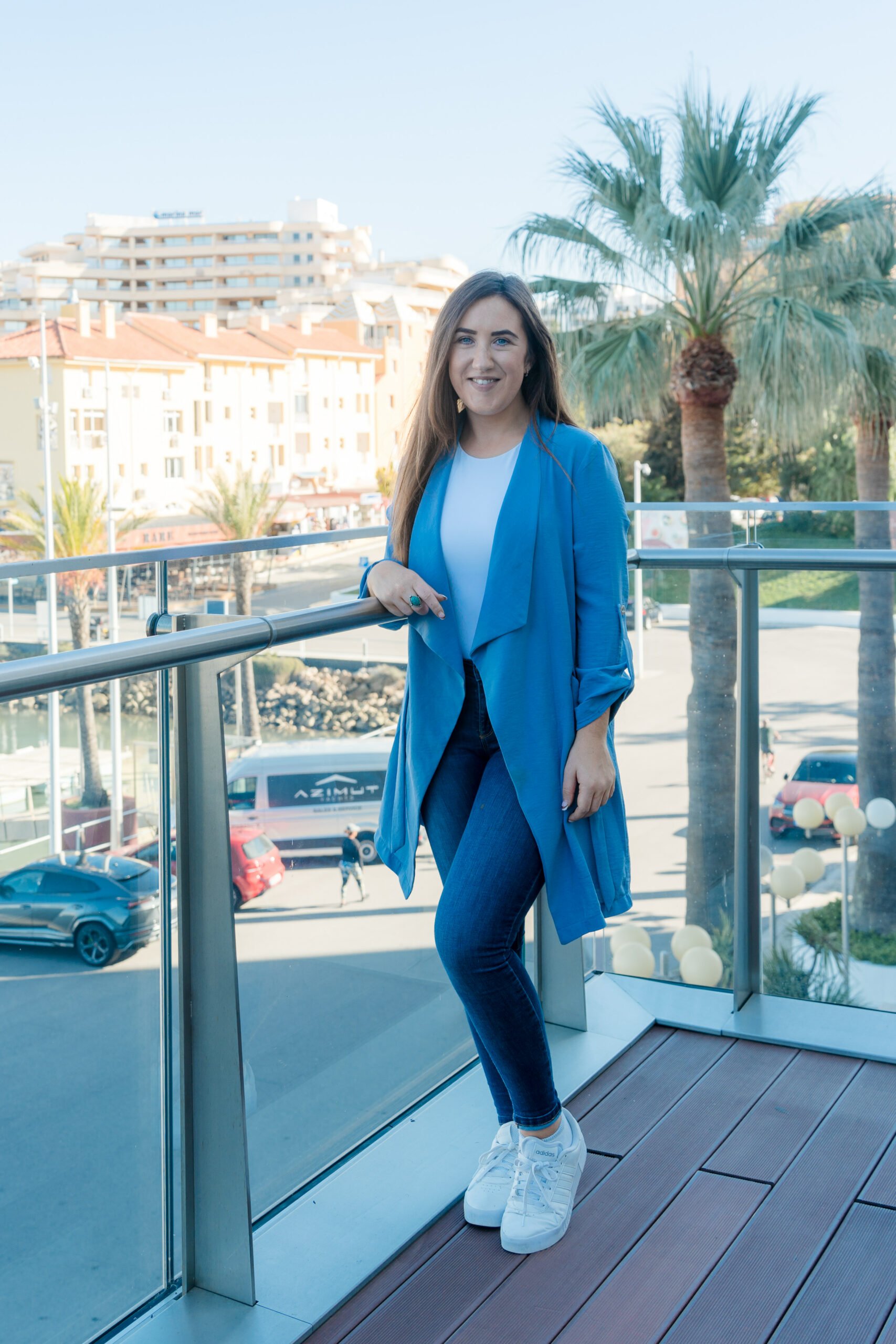 Young woman with long brown hair in a blue blazer, white shirt, and jeans, standing on a balcony with palm trees and a cityscape in the background.