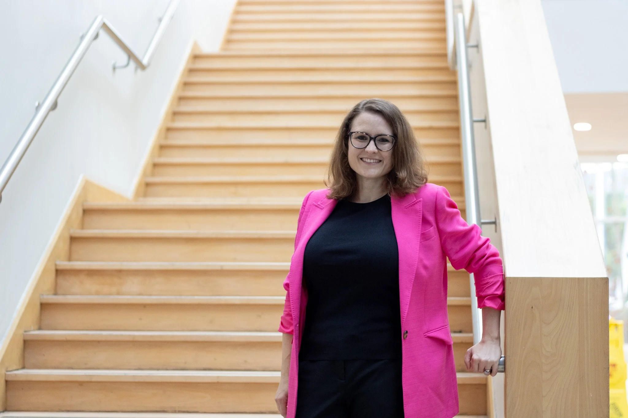 A woman with shoulder-length brown hair and glasses, wearing a pink blazer and black shirt, smiling while standing on a wooden staircase.