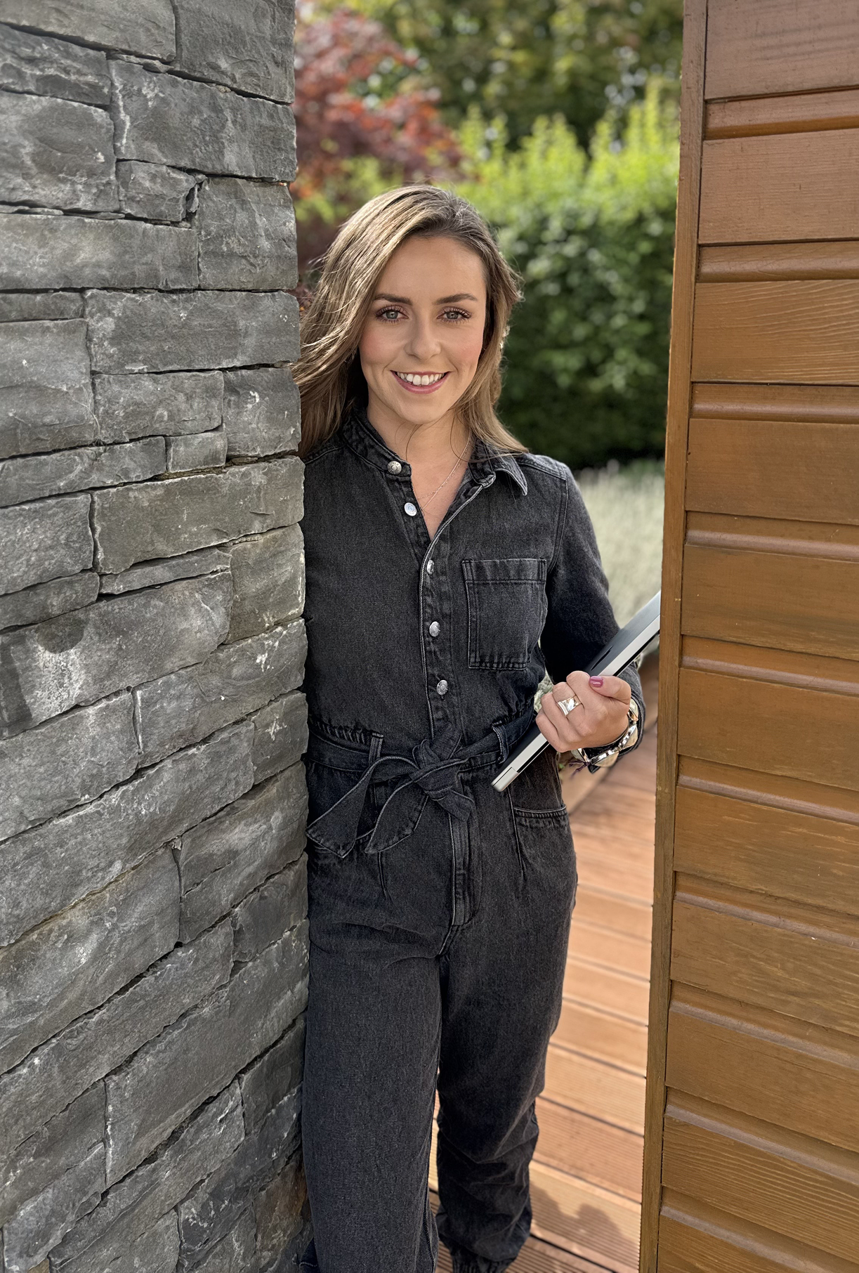 A woman smiling and standing between a stone wall and a wooden wall, holding a tablet outside with greenery in the background.