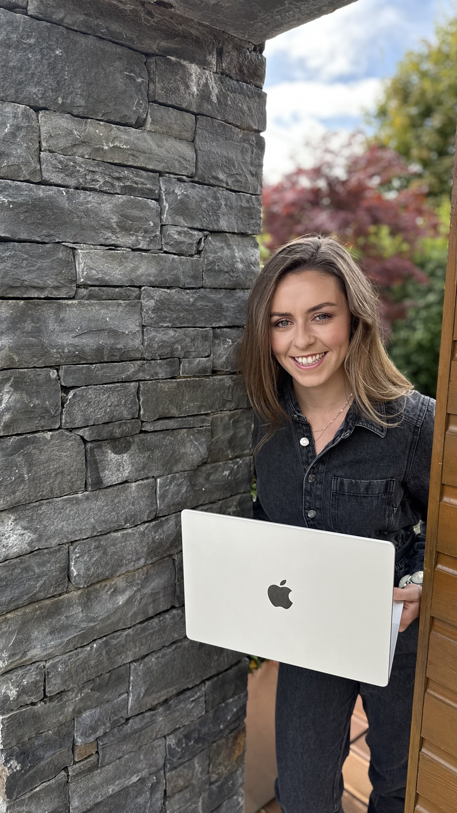 Woman with brown hair smiling and holding a MacBook laptop, standing next to a stone wall and a wooden fence outdoors.