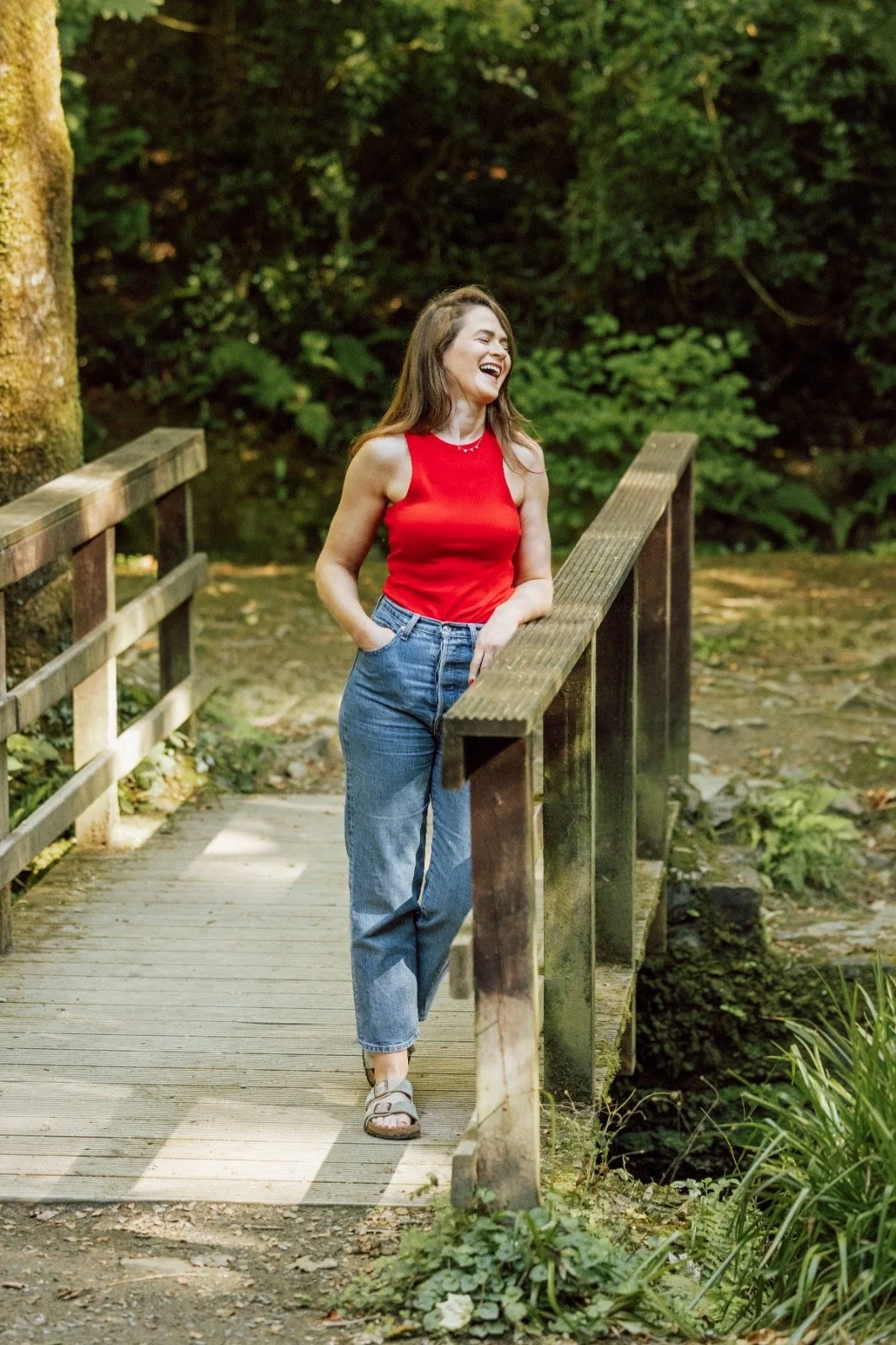 A woman with long brown hair, wearing a red sleeveless top and blue jeans, standing on a small wooden bridge in a lush green forest, laughing.