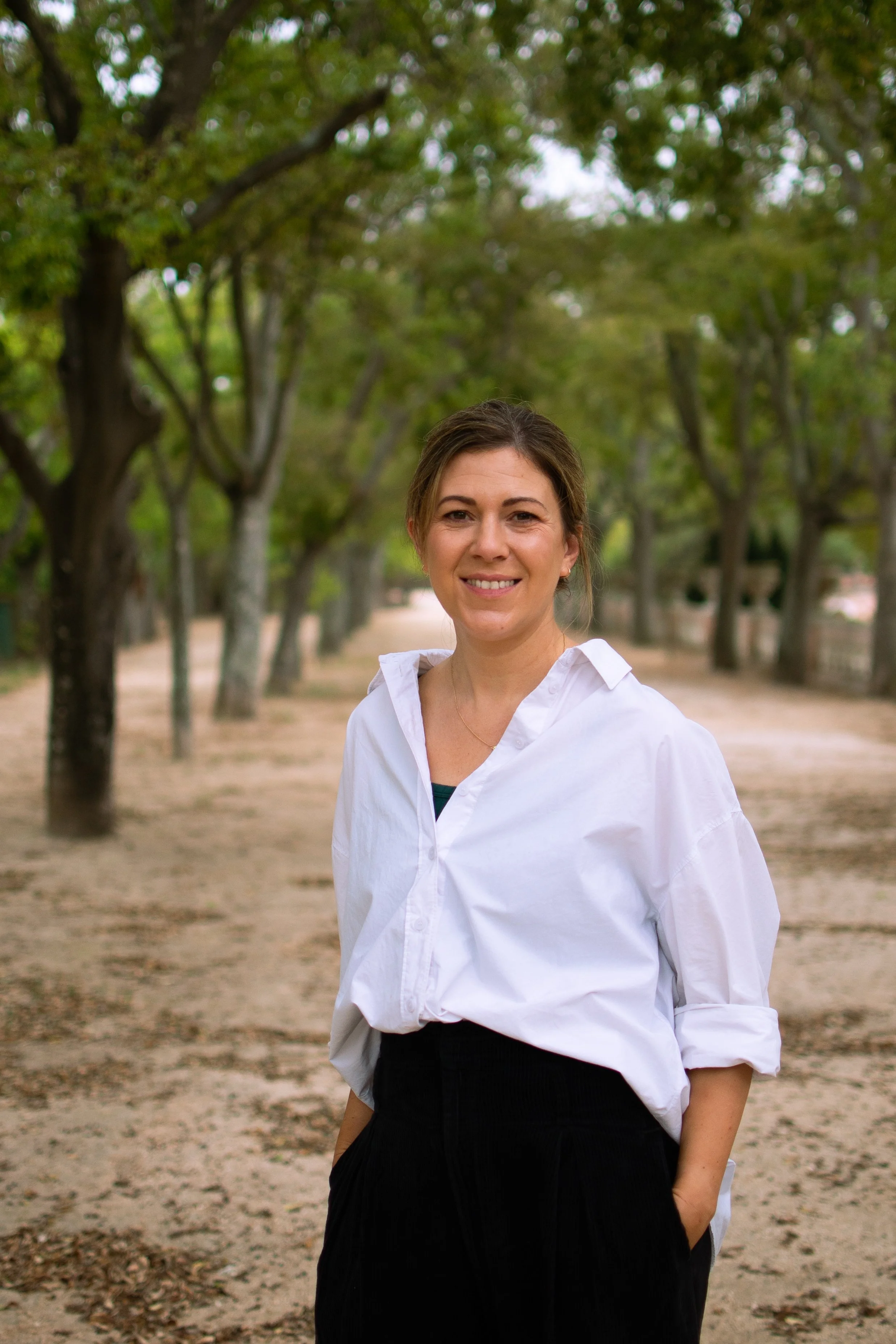 A woman with long, wavy hair sitting on a wooden windowsill indoors, with greenery outside the window, smiling at the camera.