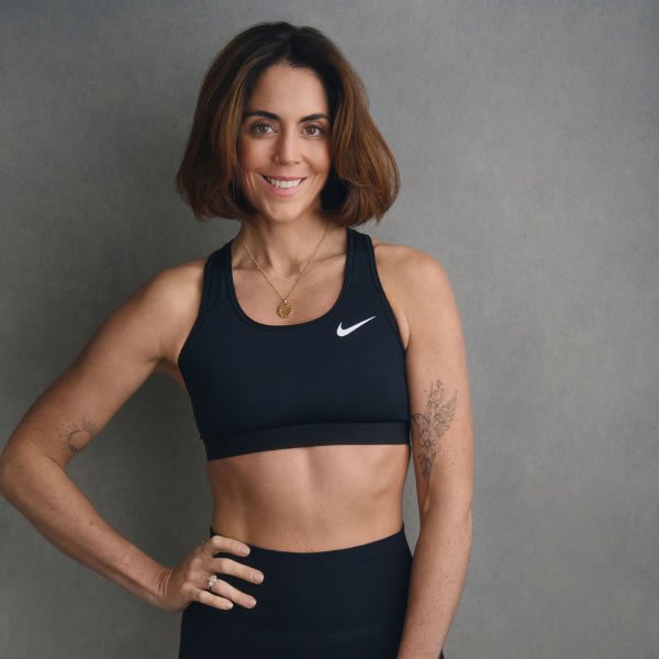 A woman with short brown hair smiles at the camera, wearing a black Nike sports bra and black leggings, standing against a gray background.