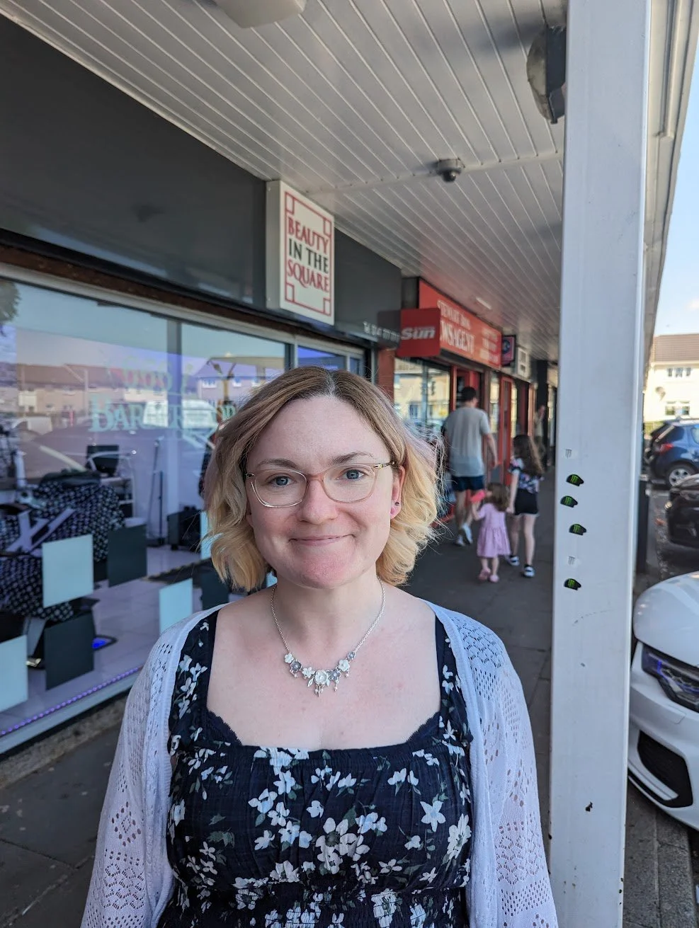 A woman with blonde hair, glasses, wearing a floral dress and white cardigan, smiling at the camera outside a storefront with signs that read 'Beauty in the Square' and 'The Sun' on a sidewalk.