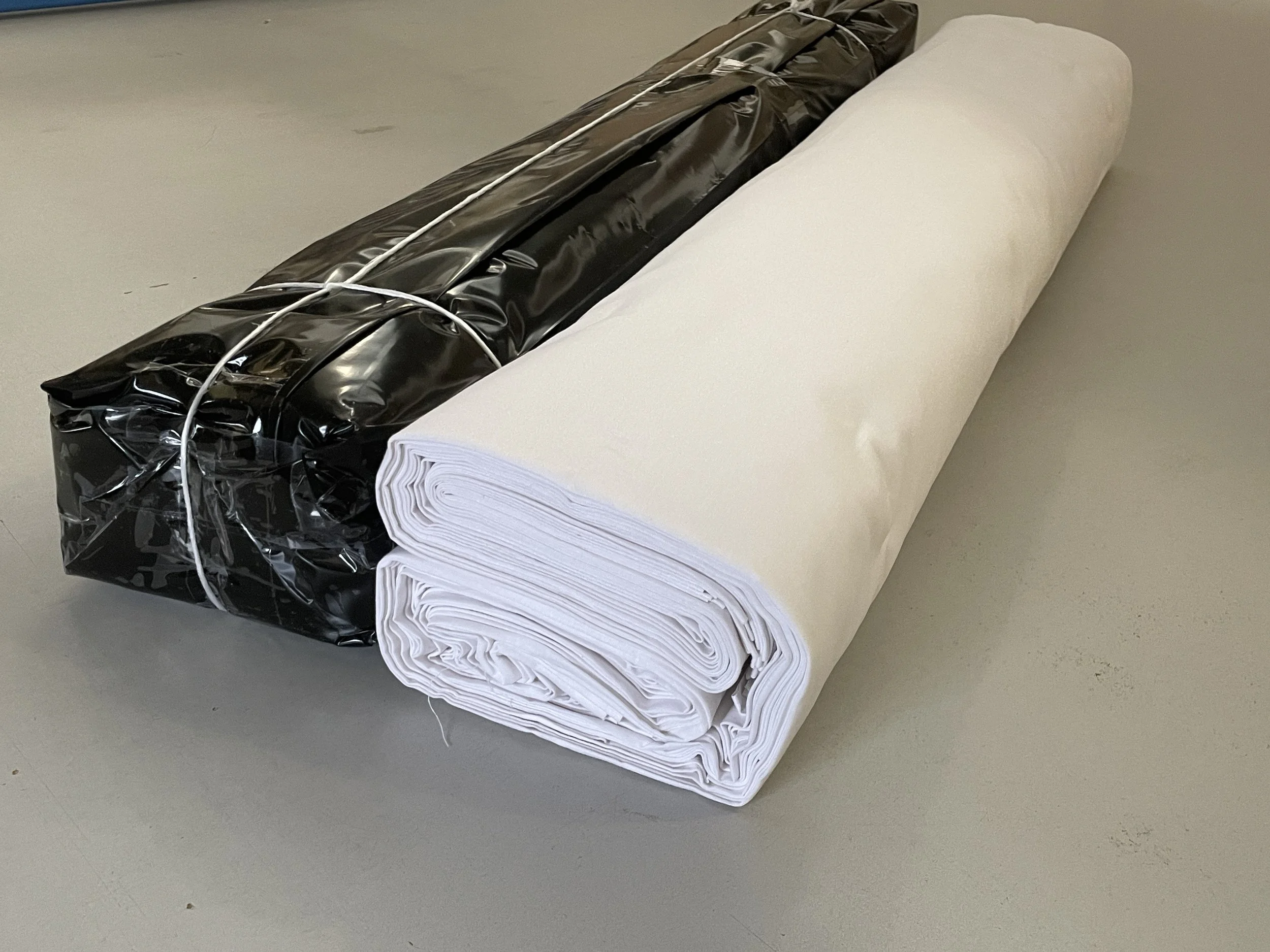A roll of white fabric or paper next to a tightly rolled black plastic sheet wrapped with elastic bands on a gray surface.