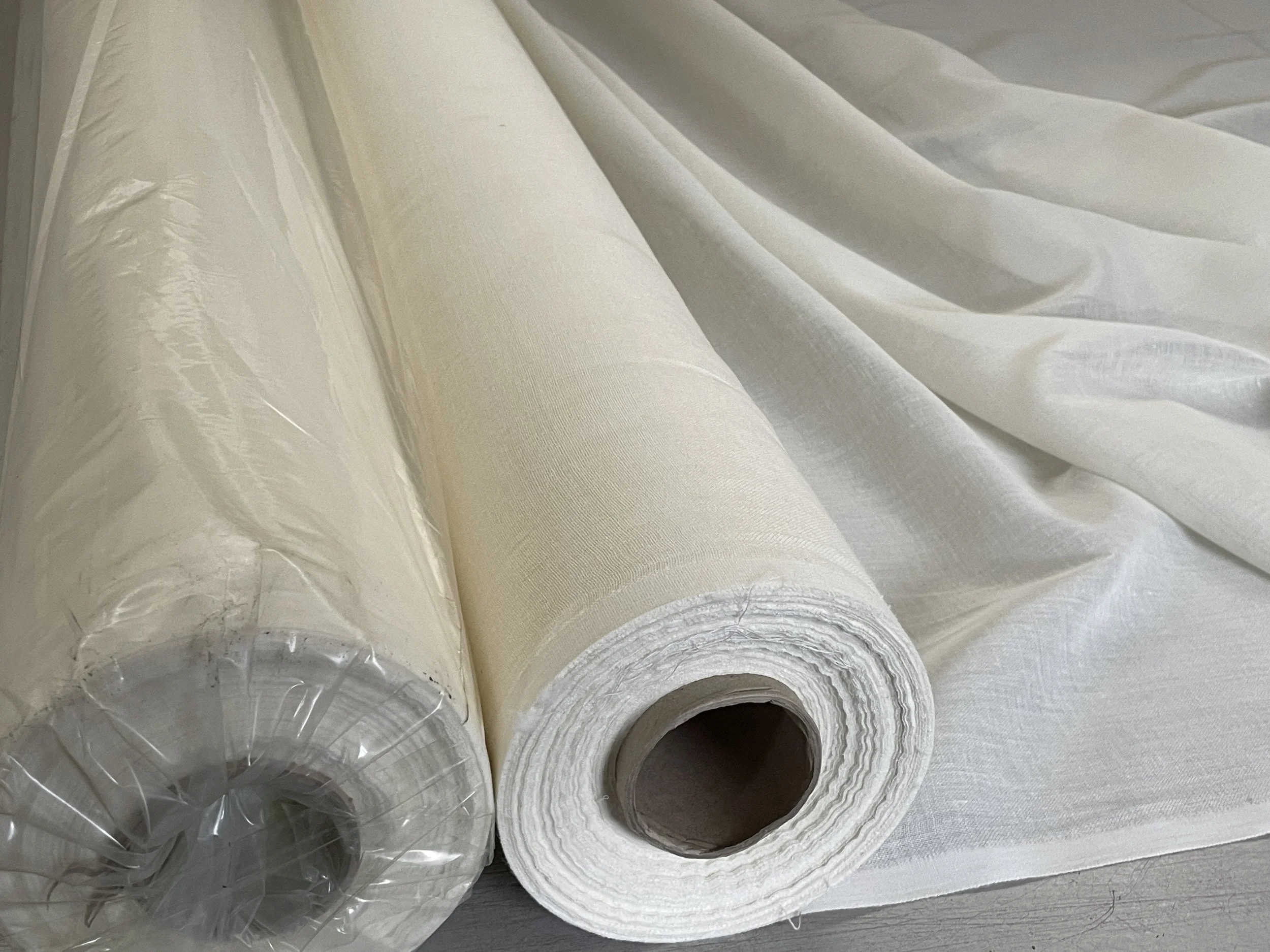 Two rolls of white fabric, one in plastic wrapping and one unwrapped, lying on a flat surface with fabric partially unfolded.
