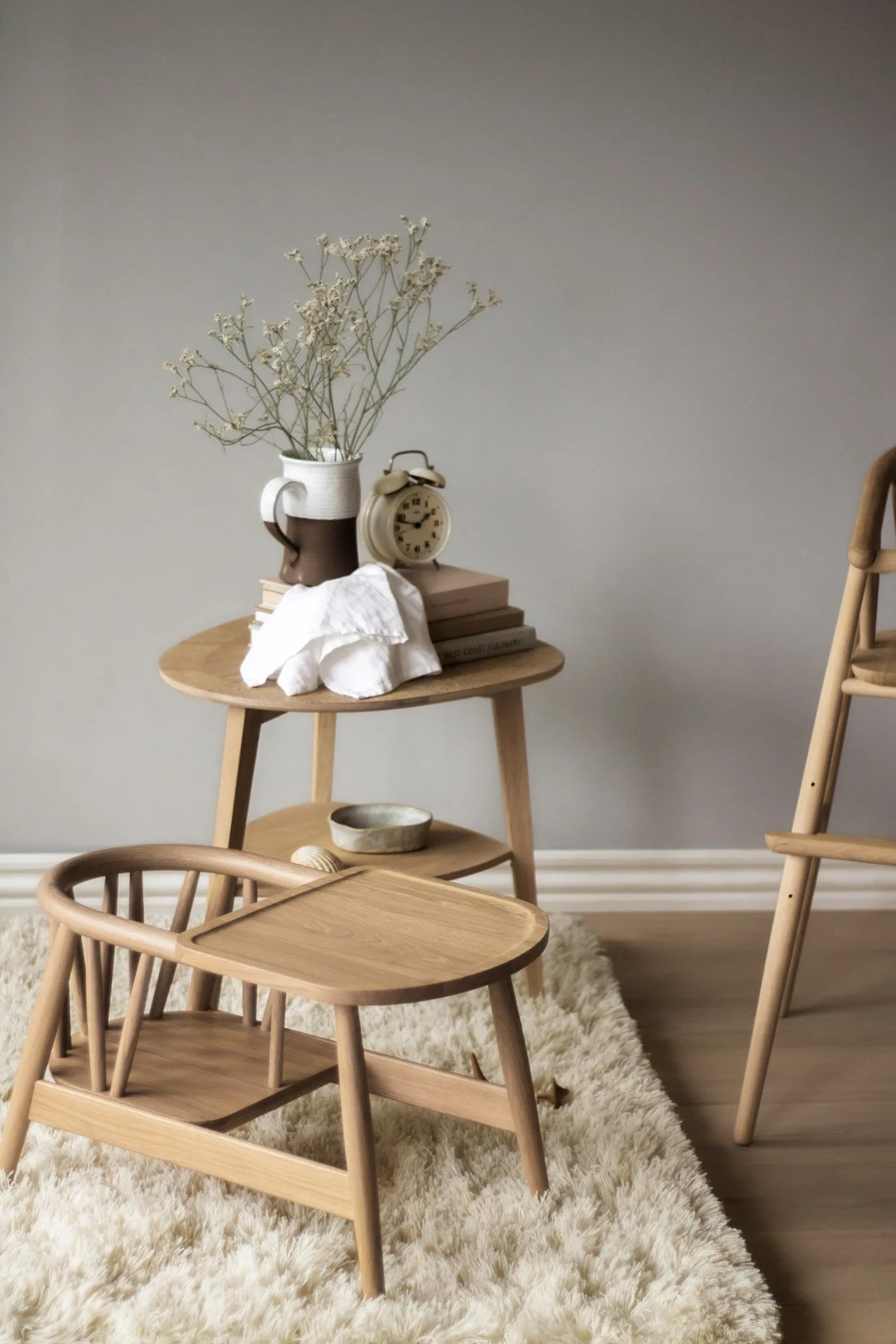 Et lille tieret bord med en keramisk kop med blomster, en analog vækkeur, flere bøger, en papirserviet og en skål står på en fluffy, cremefarvet løber. Der er også en halvt synlig træstol på højre side.