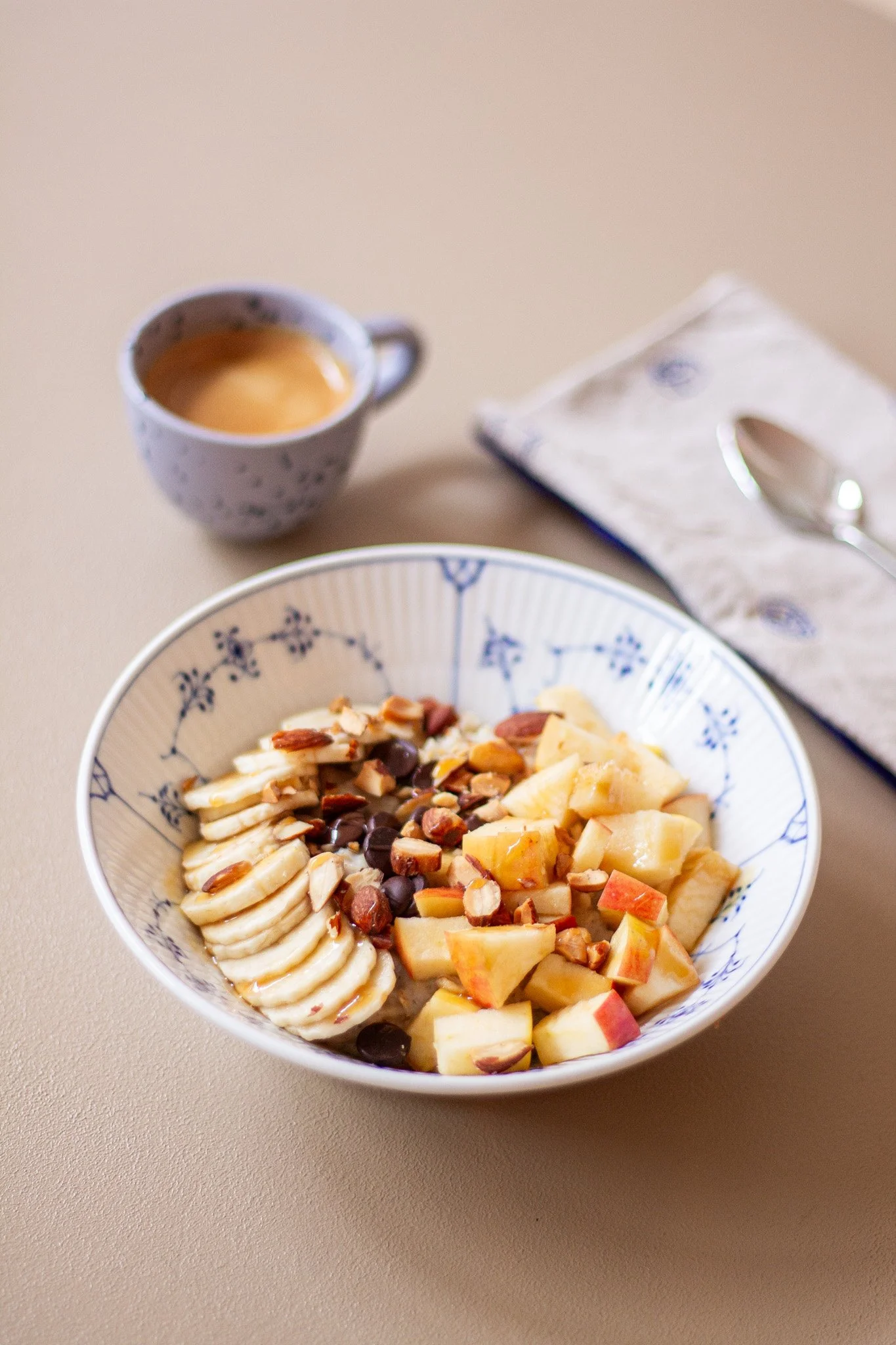 Skål med skårne bananer, æblebiter, chokoladechips og nødder. En kop med kaffe og en ske ligger på en dug med en tallerken, der er dækket af en stofserviet.