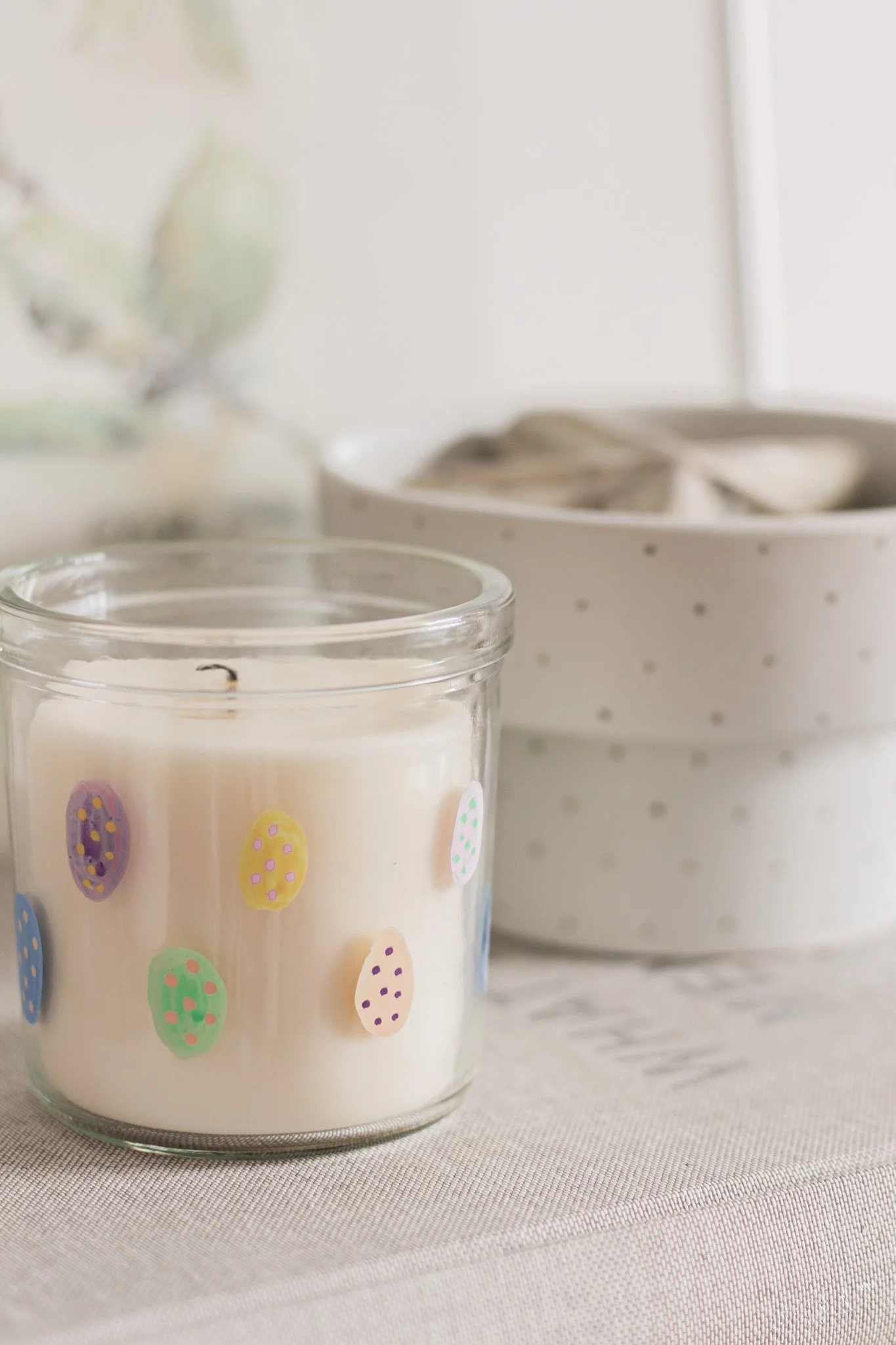 Lyse lys i en glaslysestage med ægstribende mønster og i baggrunden blå og hvide æg i kasser, dekoreret til påske.