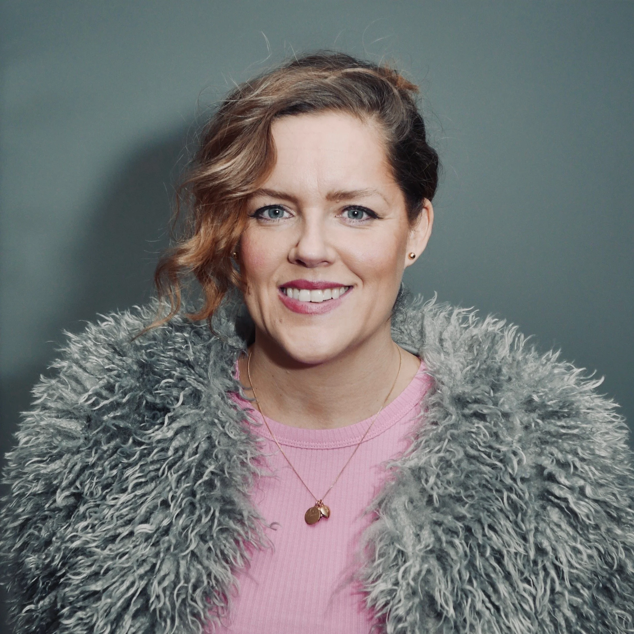 A woman with short, wavy brown hair and blue eyes, smiling at the camera, wearing a pink shirt and a gray faux fur coat, against a plain grayish background.