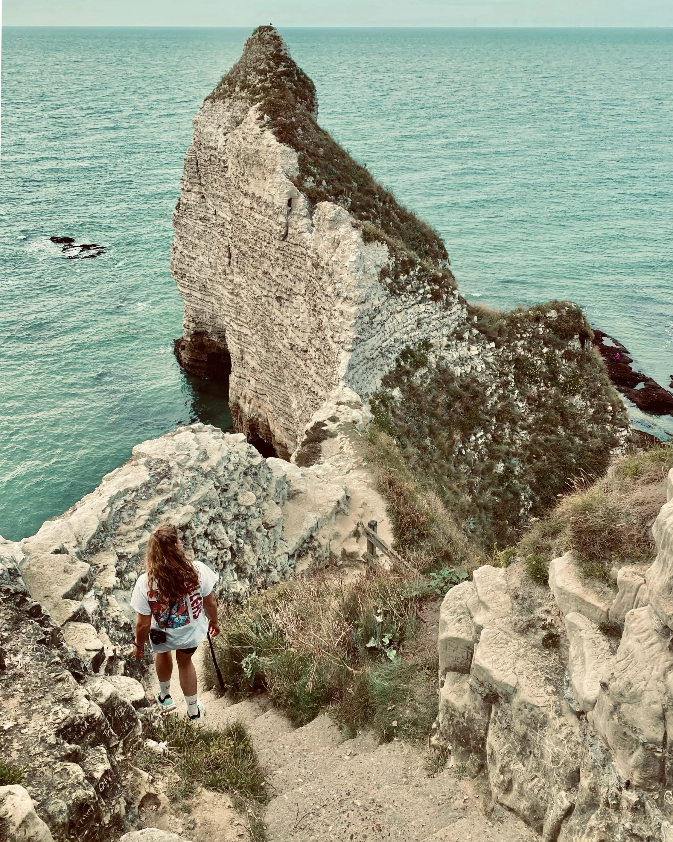 A woman with long light brown hair, wearing a white t-shirt, shorts, and carrying a backpack, walking down a rocky coastal trail with cliffs and the ocean in the background.